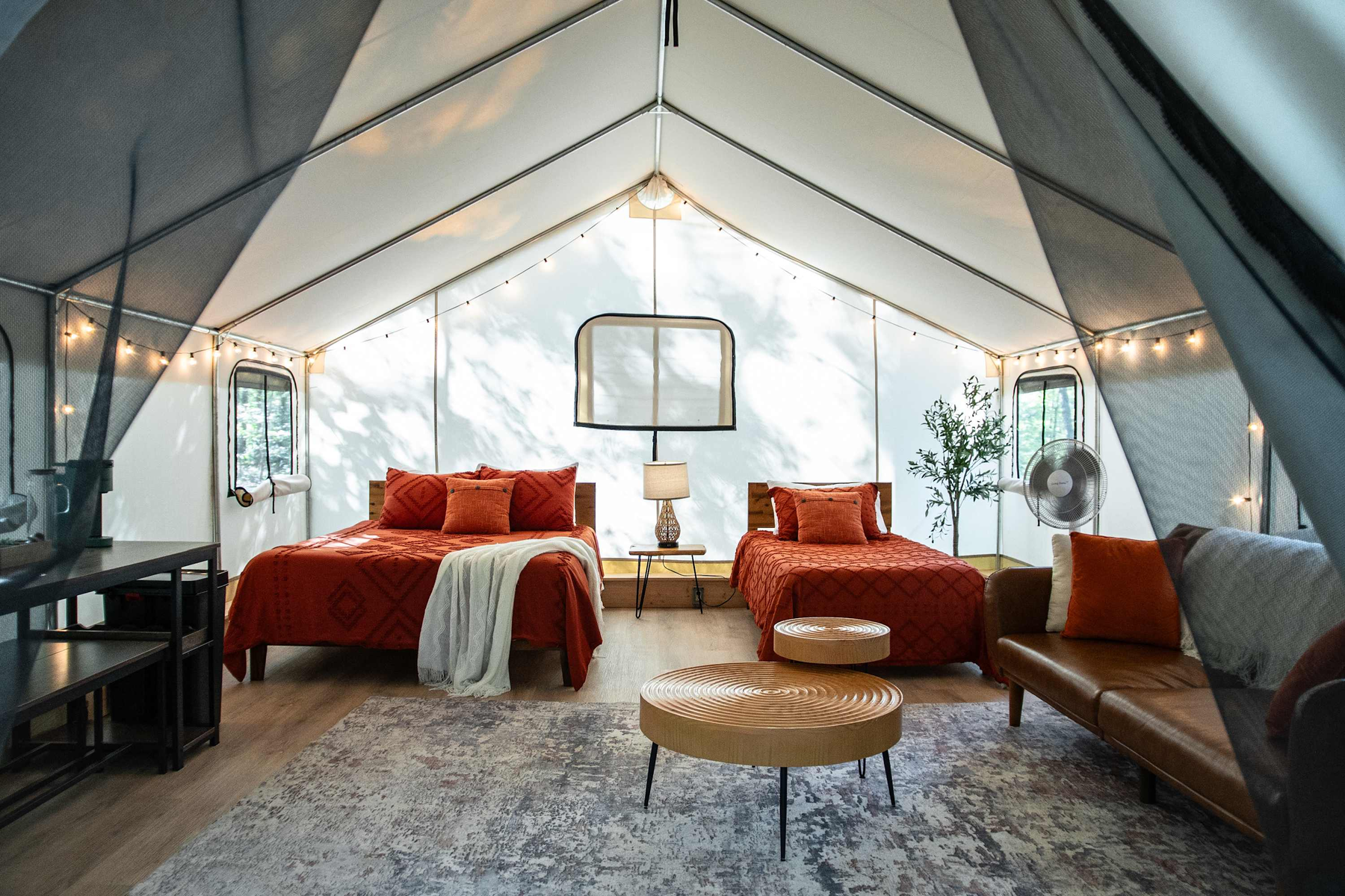 Interior of a cozy glamping tent with two beds dressed in orange bedding, a sofa, and tables, decorated with string lights and a rug, with windows and a fan.