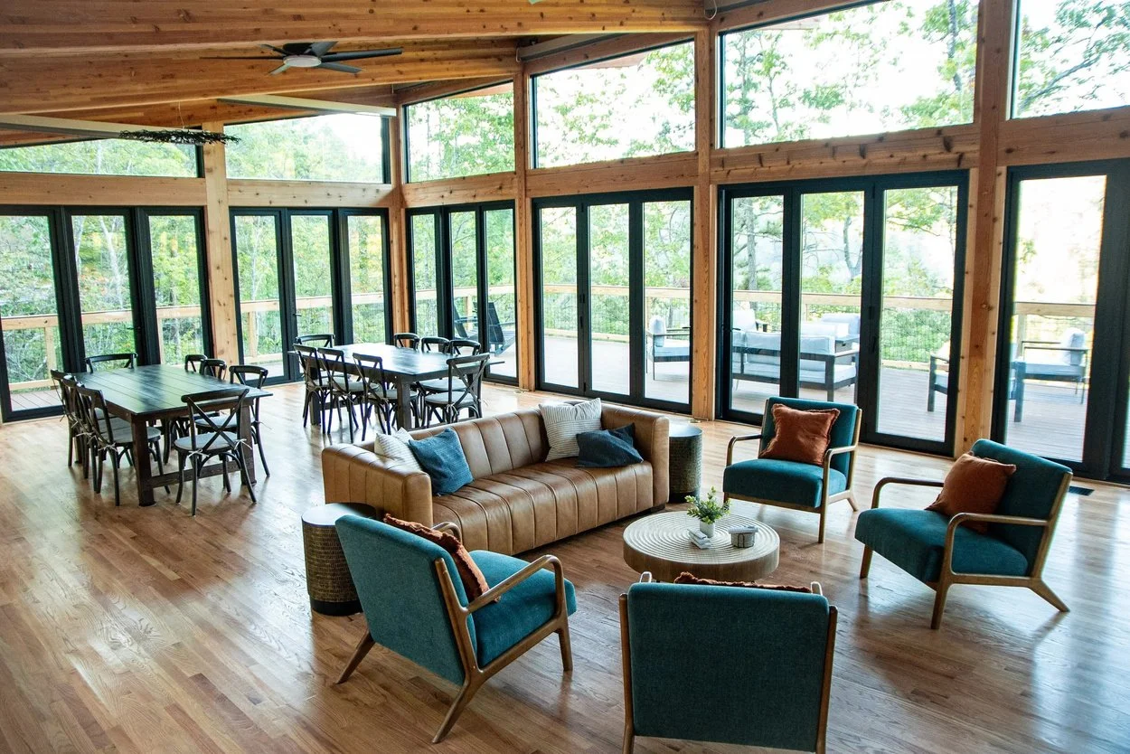 Living room with wood floors, large glass windows, a tan leather sofa, four teal armchairs, a round wooden coffee table, and a dining table with ten chairs. Outside deck with seating is visible through the windows.