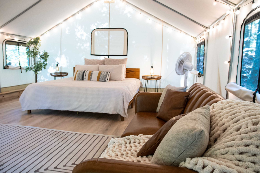 Inside a cozy tent with a bed and a brown sofa, decorated with string lights, pillows, a mirror behind the bed, a fan, and windows showing trees outside.