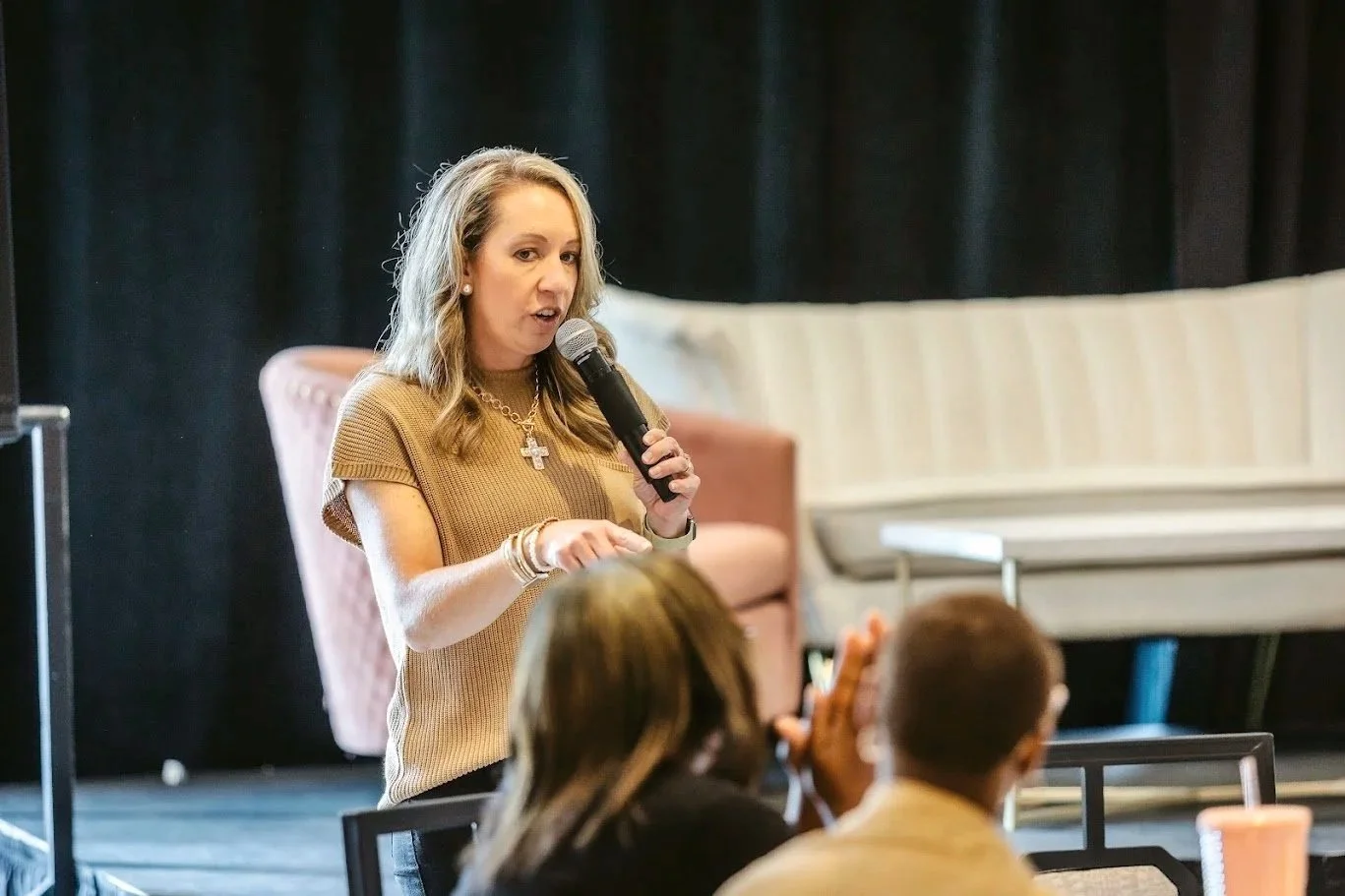 A woman with blonde hair holding a microphone, speaking to an audience in a room with a piano and black curtains.