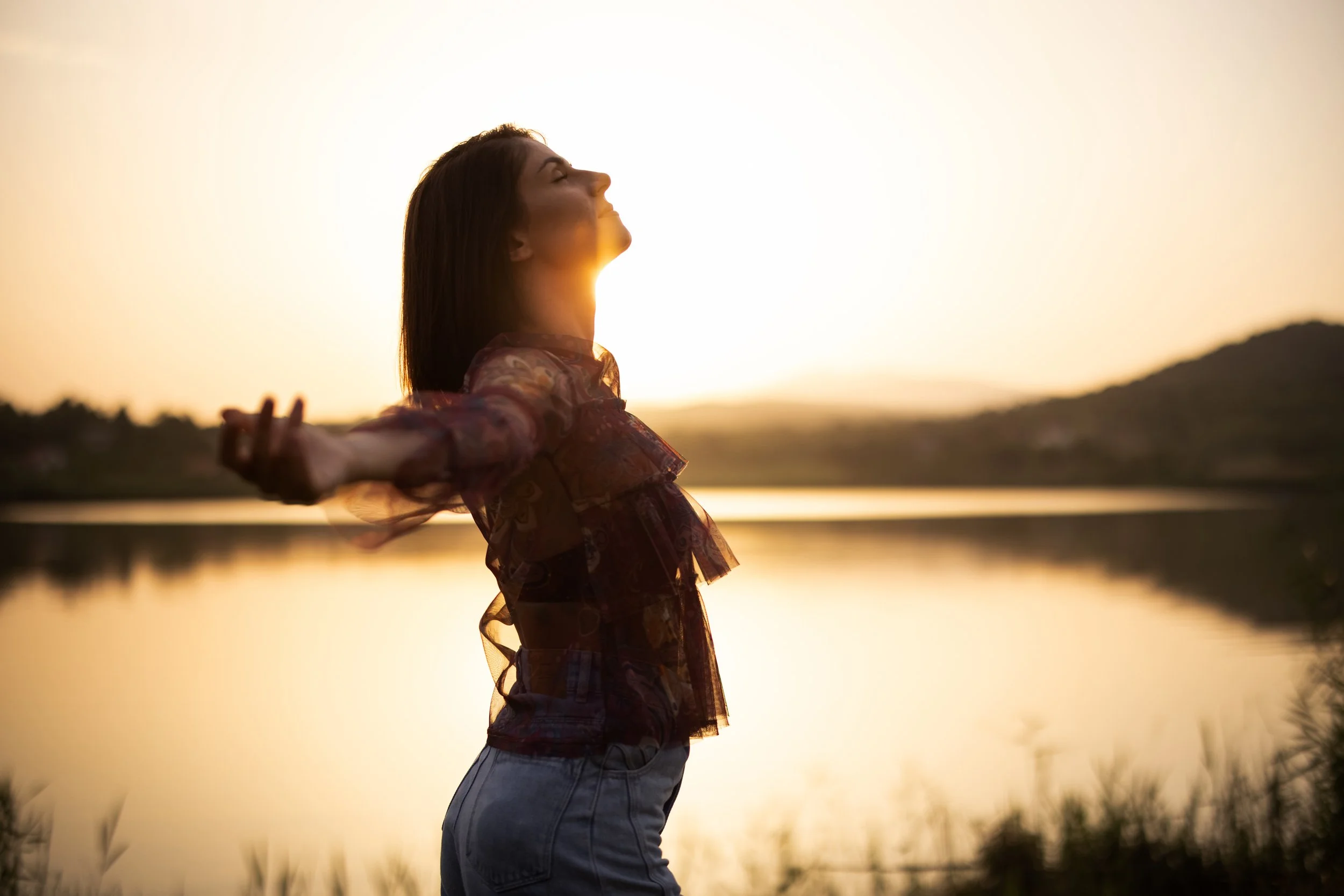 Woman standing by a lake at sunset with arms outstretched and face lifted, enjoying the moment.