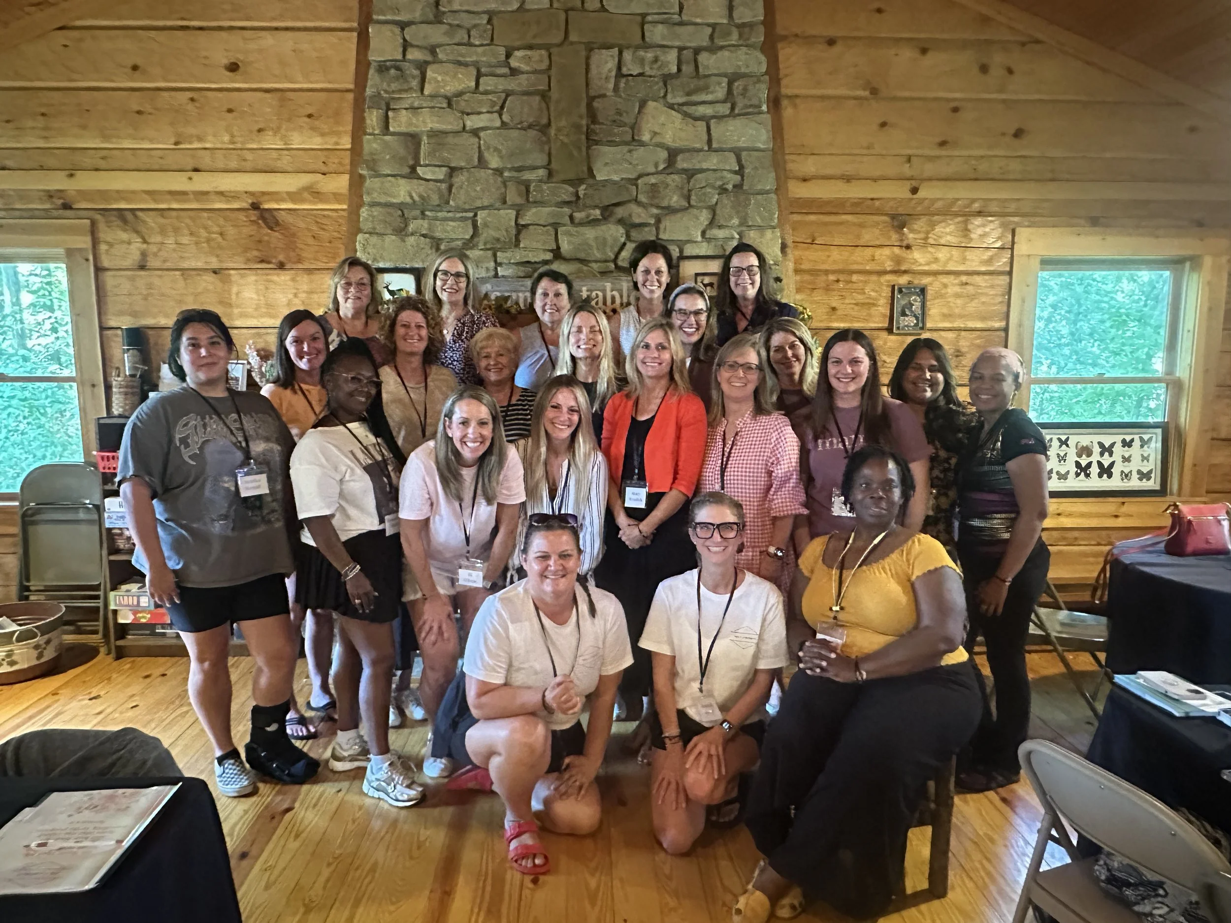 Group photo of women in a wooden cabin with a stone fireplace, some standing and some sitting, smiling at the camera.