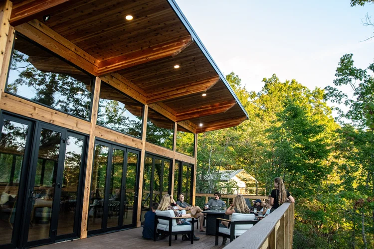 People gathering and socializing on a wooden deck overlooking trees, with large glass windows and a wooden ceiling on the house.