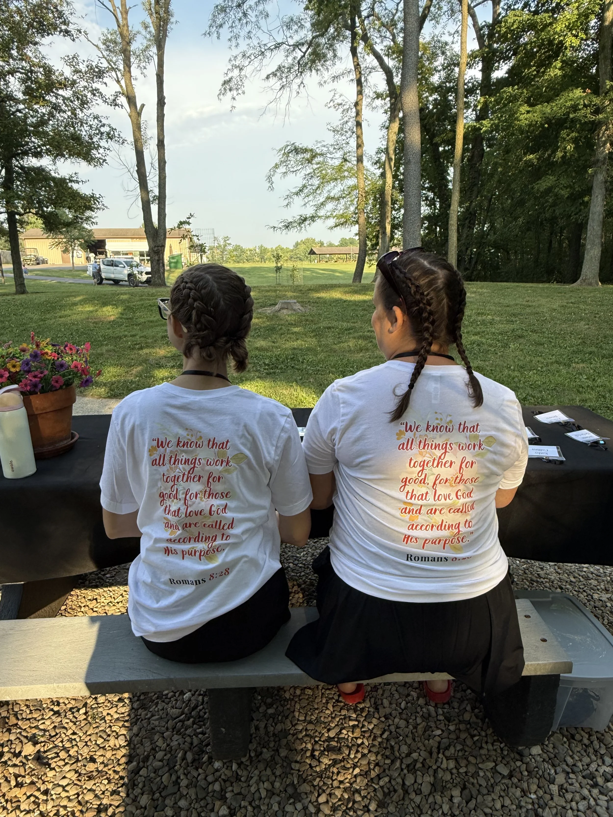 Two women with braided hair and glasses sitting on a park bench at a table, wearing white t-shirts with a Bible verse printed on the back, outdoors in a park with trees and a grassy area.