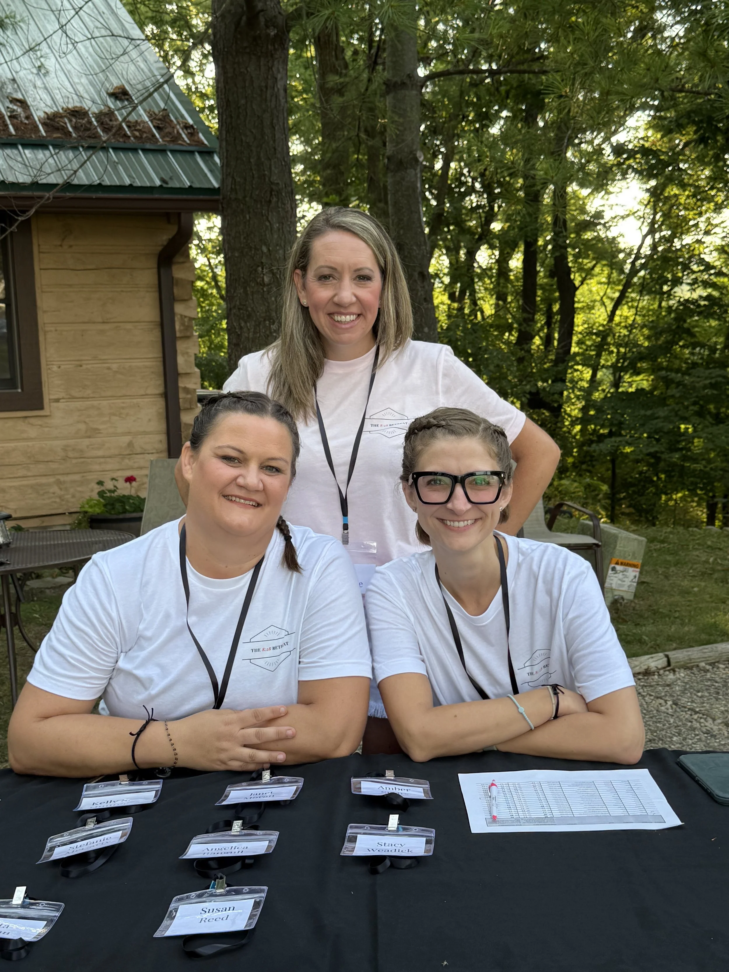 Three women wearing matching white T-shirts and black lanyards are sitting and standing at an outdoor table with name tags and sheets, with trees and a wooden building in the background.