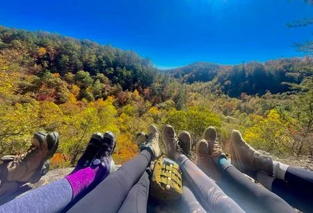 Six people sitting on a ledge overlooking a forested mountain landscape on a clear day