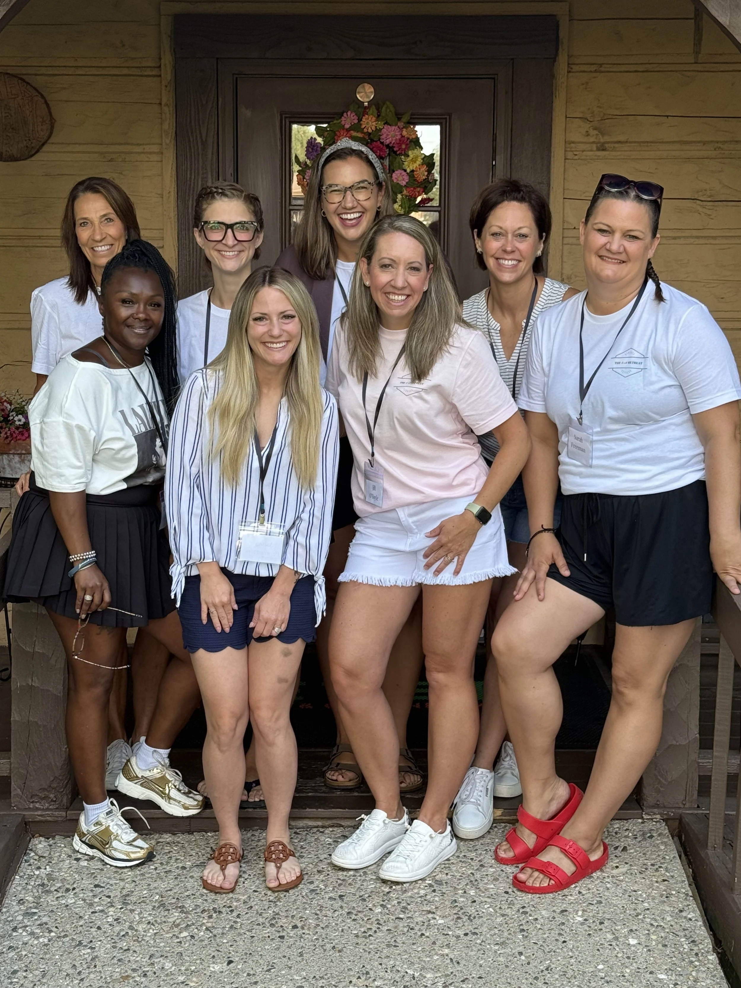 Group of nine women standing on porch in front of a door with a floral wreath, smiling at camera.