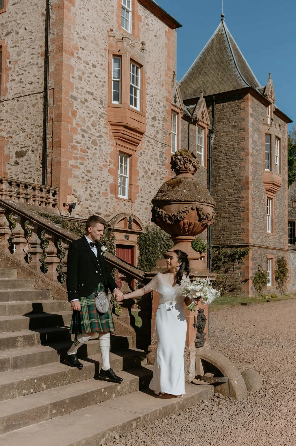 A bride and groom holding hands on stone stairs outside a historic mansion. The bride is wearing a white wedding dress and holding a bouquet of flowers, the groom is wearing a traditional Scottish kilt with a tuxedo jacket. The setting is sunny with clear blue skies.
