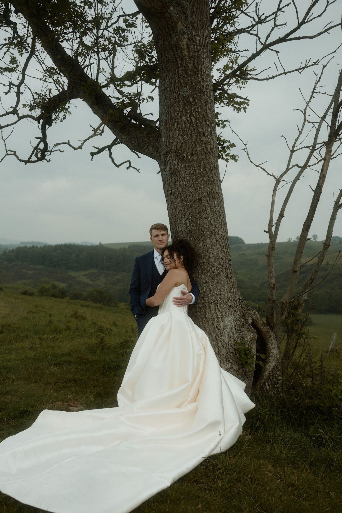 A bride and groom standing close together outdoors next to a large tree, with a scenic landscape of rolling hills in the background. The bride is wearing a white wedding gown and the groom is in a navy suit. The bride is resting her head on the groom's shoulder, and they are embracing.