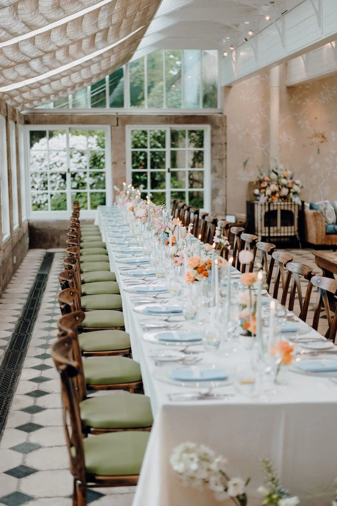 Long dining table set for a celebration with pink and orange floral arrangements and candles, in a bright room with large windows and green foliage outside.