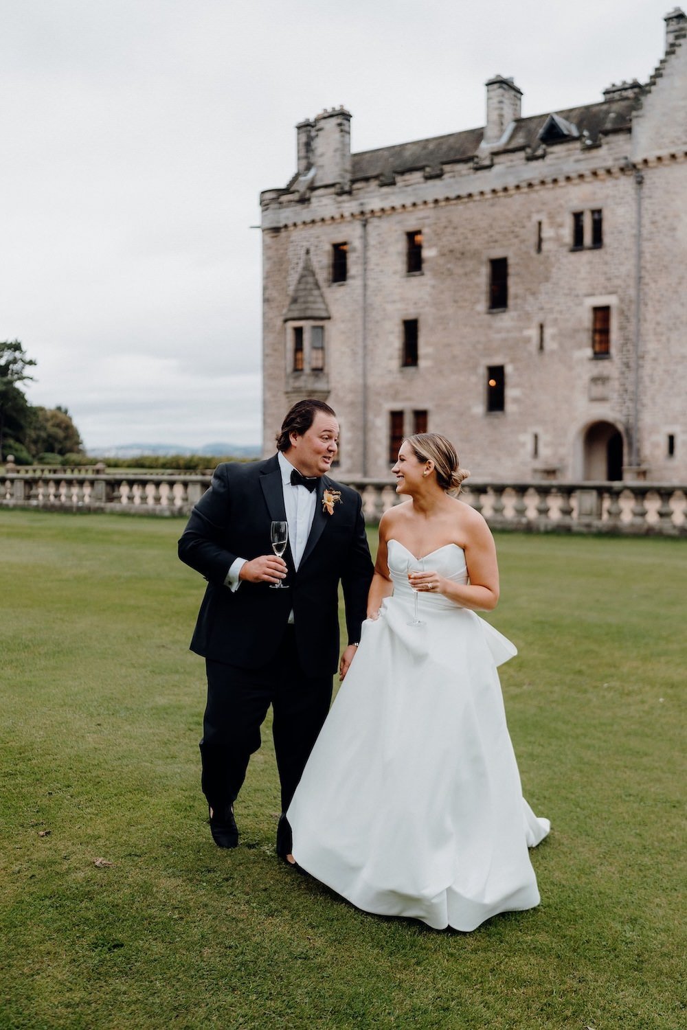 A newlywed couple at Banrbougle Castle with glasses of champagne walking on a lawn in front of a castle, smiling and enjoying their wedding day.