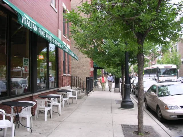 Sidewalk view with outdoor seating, trees, parked cars, and pedestrians in front of an Italian wine and food shop.