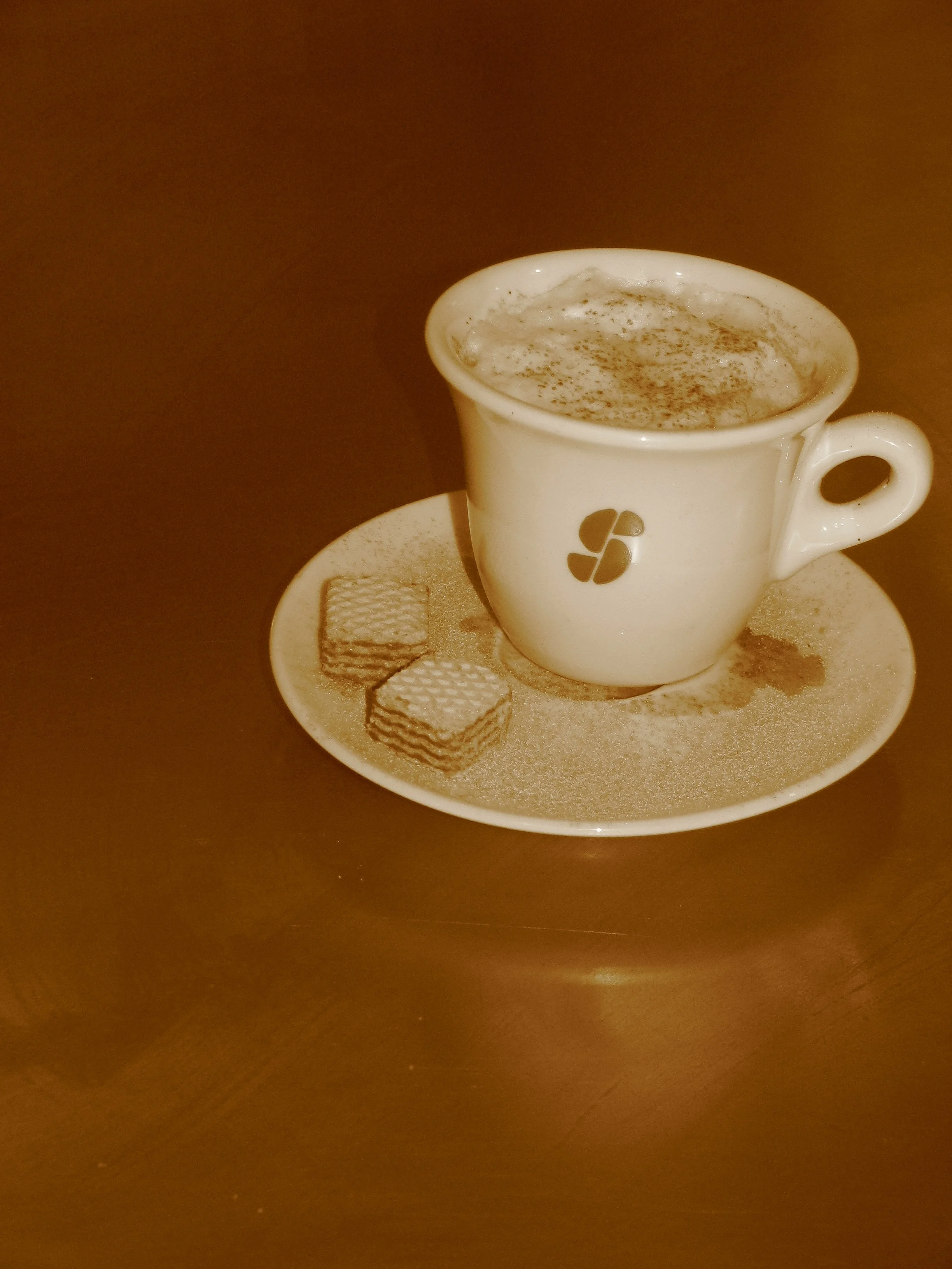 A white coffee cup filled with frothy coffee on a matching saucer, with two square biscuits on the saucer, all dusted with cocoa powder, on a dark wooden surface.