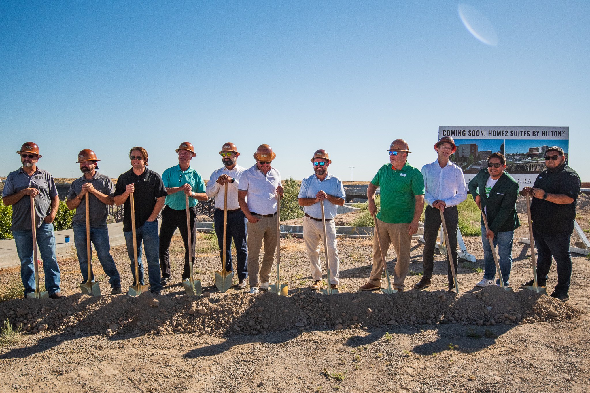 A group of people at a groundbreaking ceremony, standing in a line with shovels in hand, wearing hard hats, with a sign in the background that reads 'Coming Soon! Home2 Suites by Hilton.'