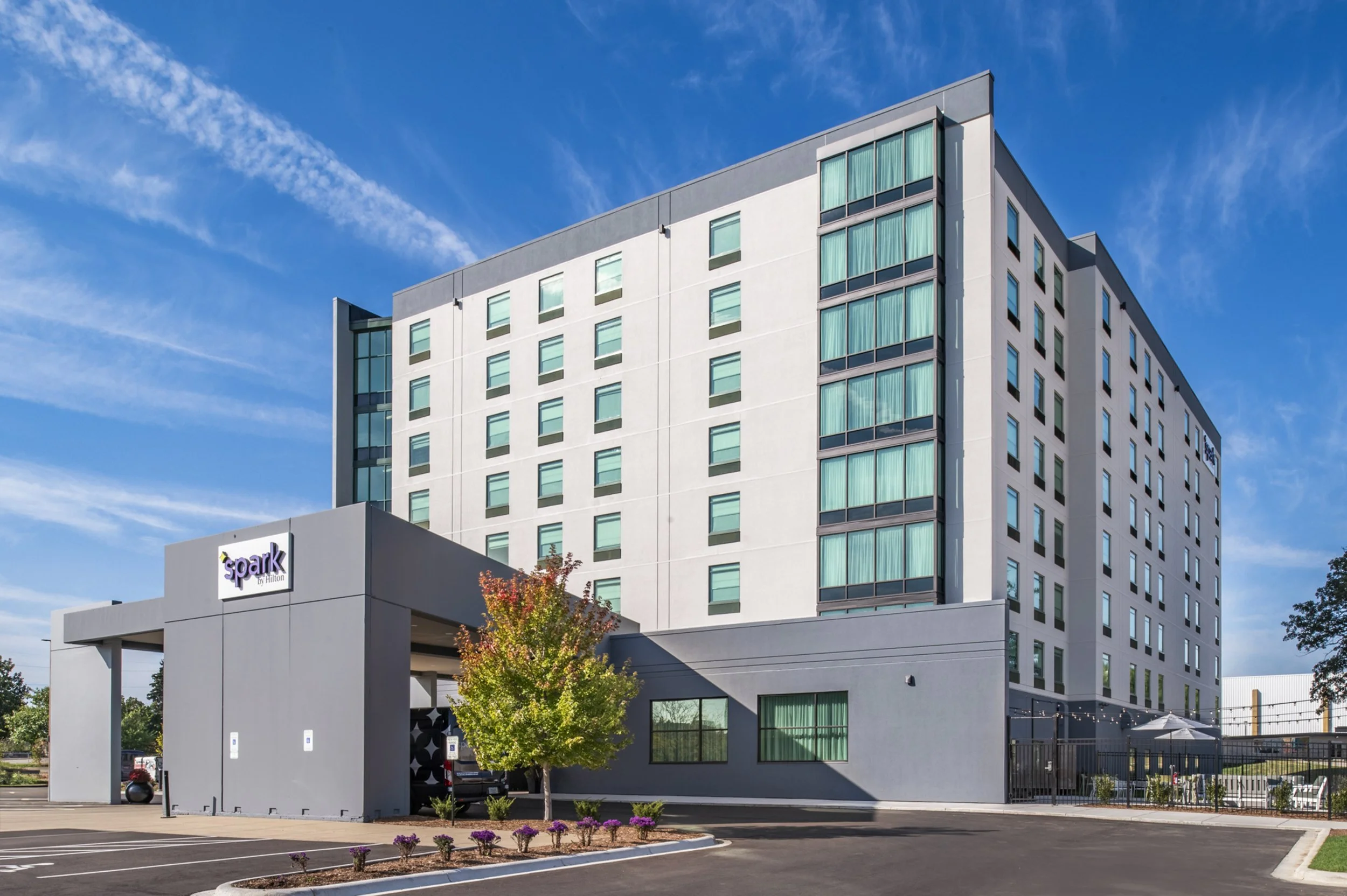 A modern multi-story hotel building with a sign that says 'Spark by Hilton,' featuring a parking lot with a few cars and a small landscaped area with trees and flowers.