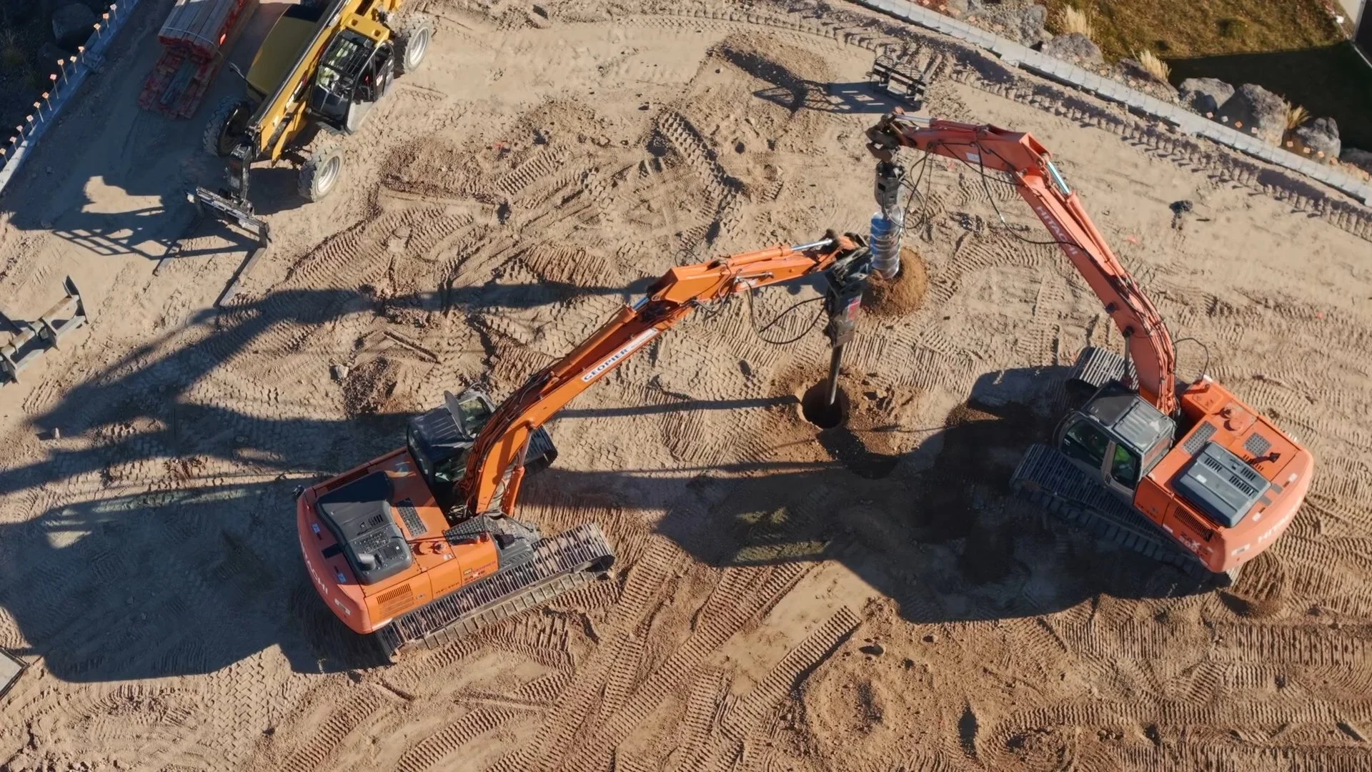 An aerial view of a construction site with two large orange excavators digging a hole, surrounded by dirt and construction equipment.