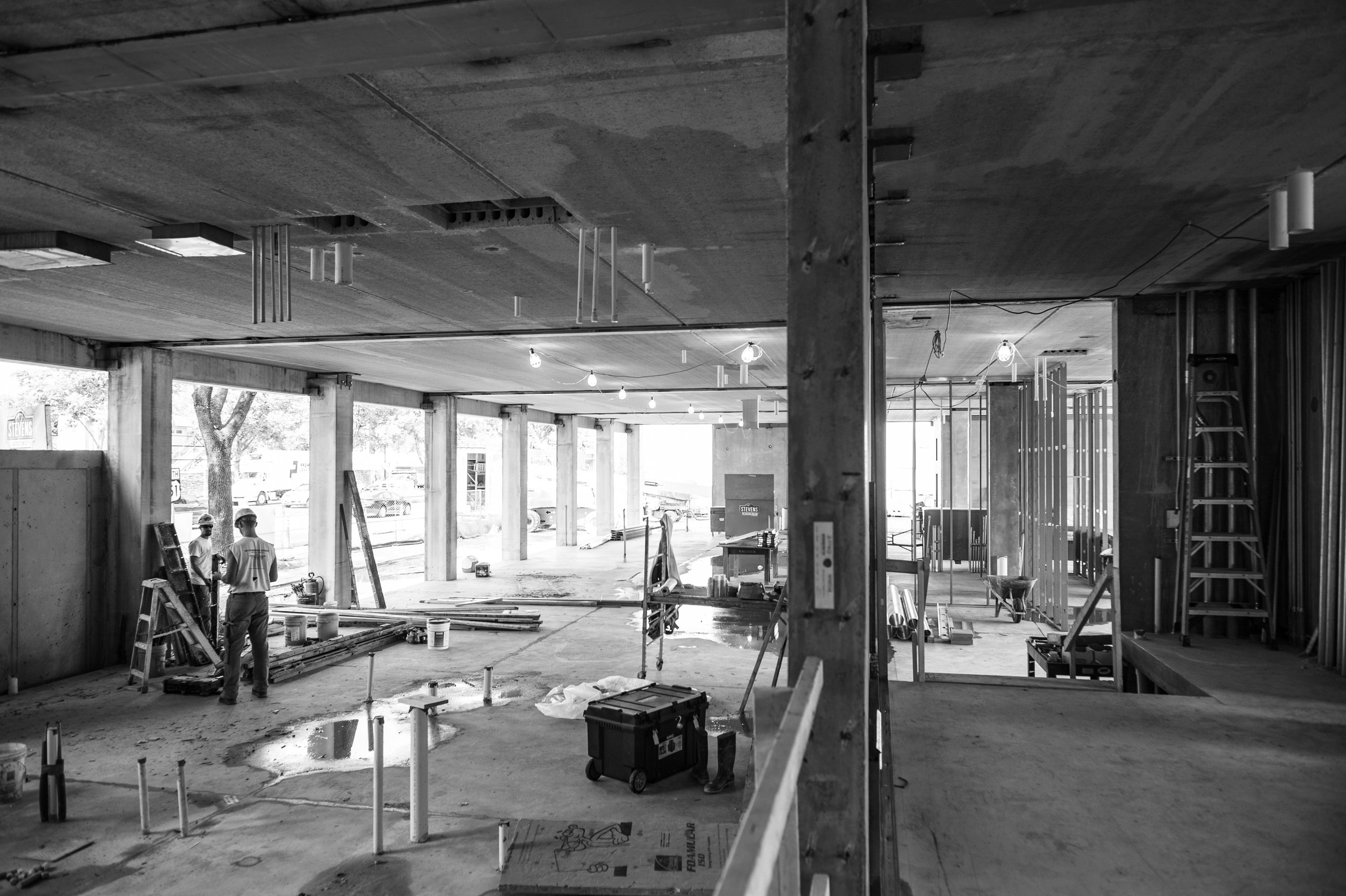 Construction workers on site inside a building under construction with exposed concrete walls and ceiling, pipes and construction tools visible.