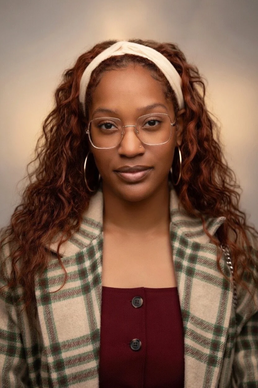 A woman with curly hair wearing a white headband, glasses, large hoop earrings, a plaid jacket, and a maroon top.