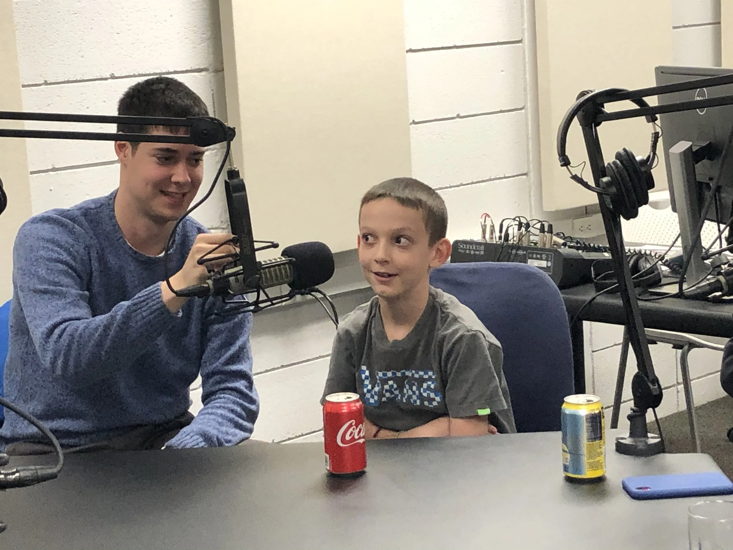 Two boys sitting at a table in a recording studio, with microphones and audio equipment. One boy is smiling, the other looking amused. There are soda cans and a phone on the table.