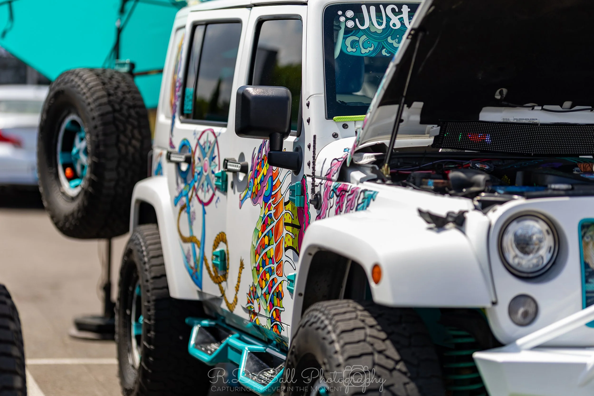 A white Jeep with colorful, psychedelic artwork on its side, open hood revealing engine, and spare tire mounted on the back.