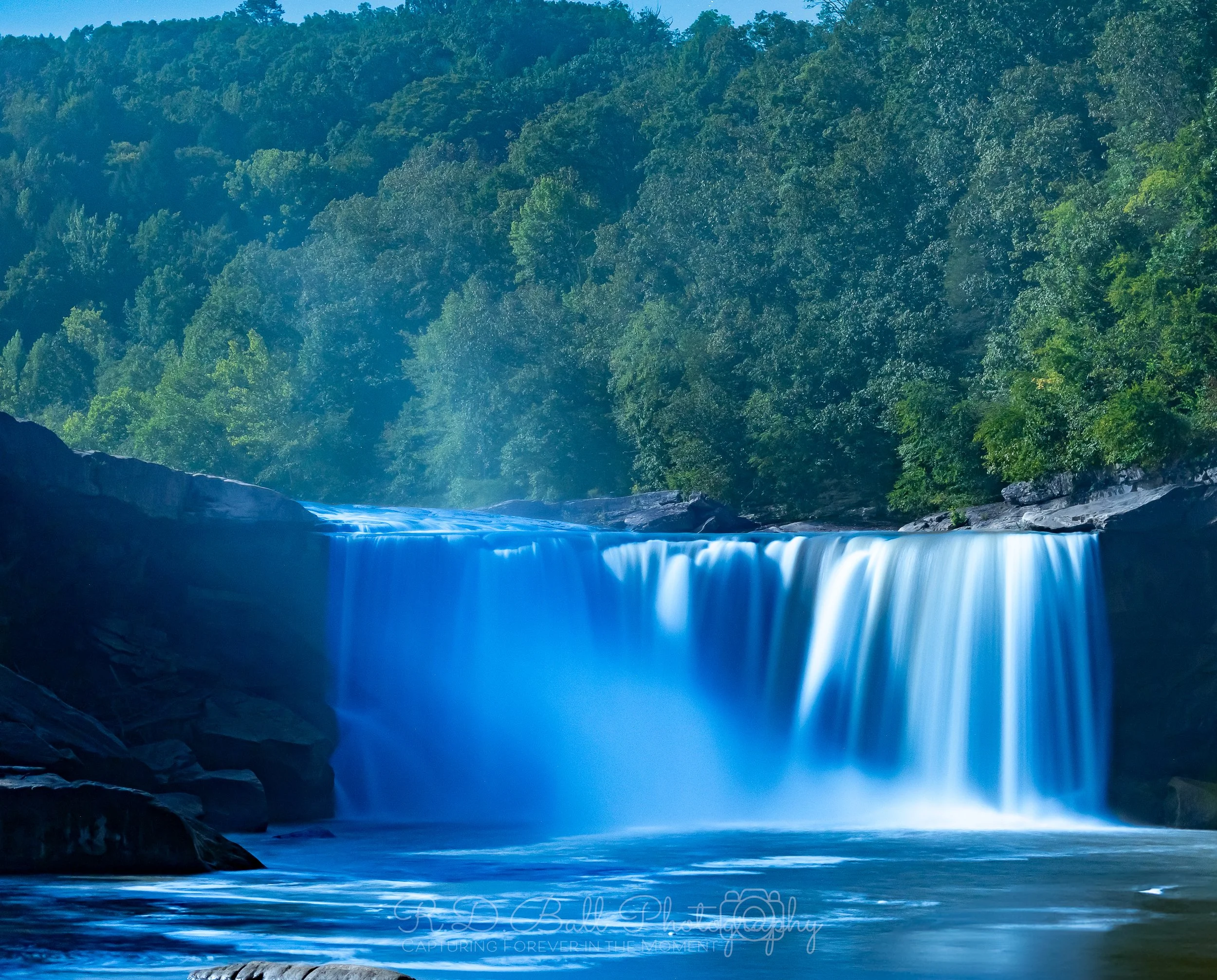 A waterfall flowing over rocks into a river, surrounded by dense green trees and hills.