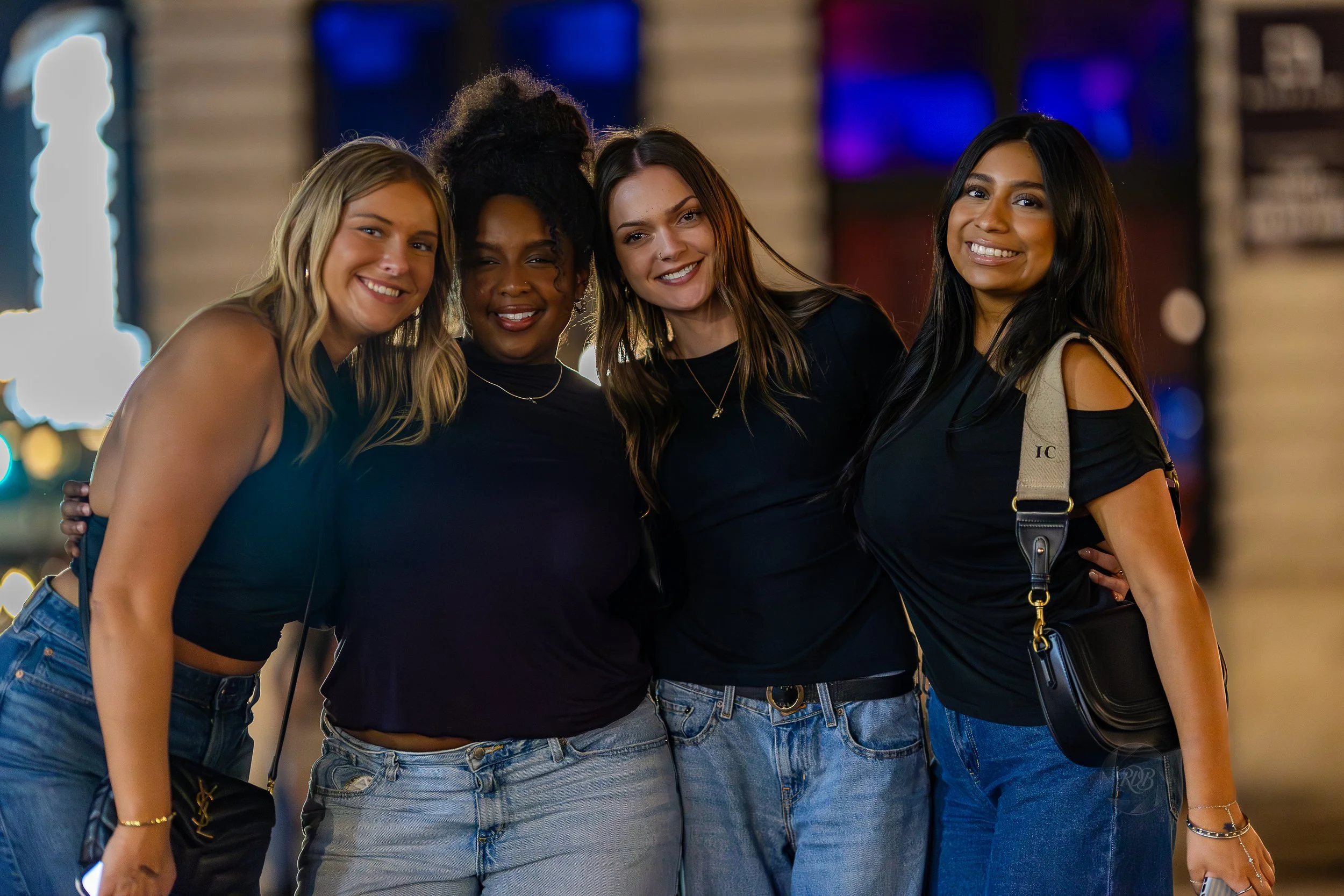Four women smiling together outside at night, dressed casually in black tops and jeans, with a blurred city background.
