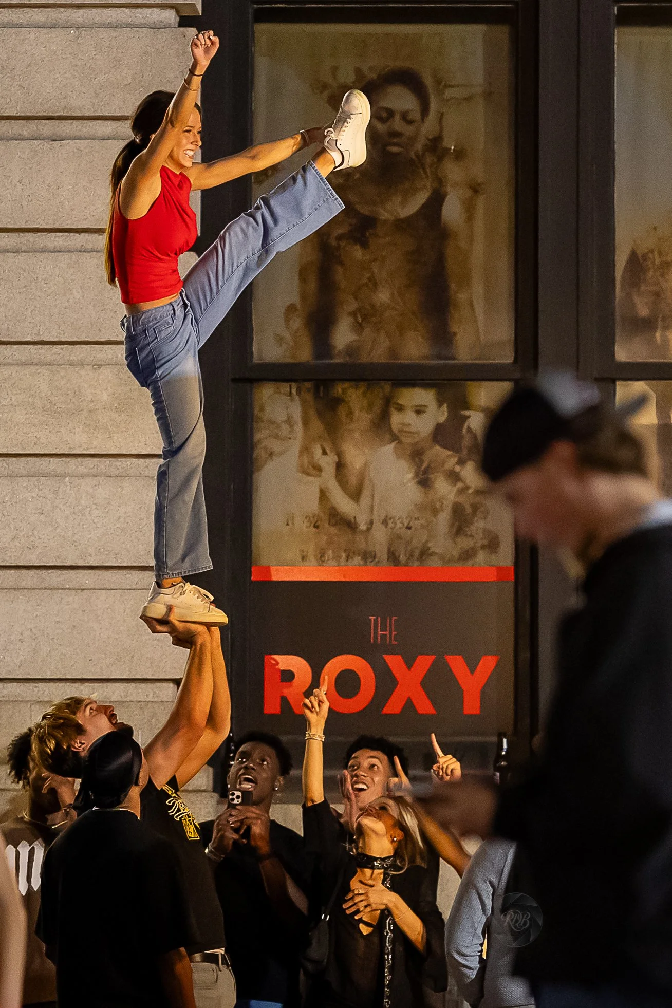 A woman in a red top and blue jeans is balanced on a man's hand, performing a cheerleading stunt called a 'liberty' during a night out in front of a poster for The Roxy. The group of women is cheering and pointing up at her.