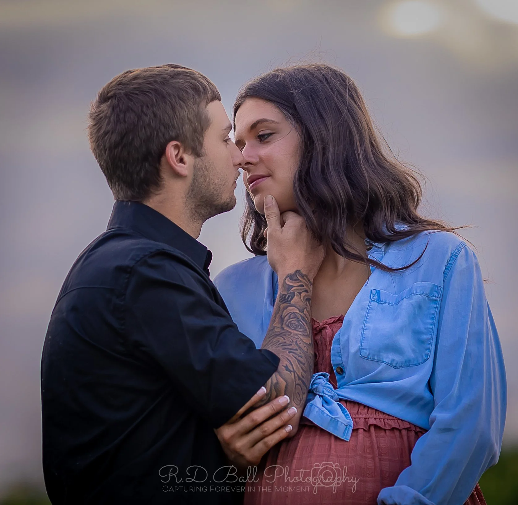 A man and woman standing close together, about to kiss outdoors during sunset. The man has short brown hair and a tattoo on his arm, while the woman has shoulder-length wavy dark hair and wears a blue shirt.