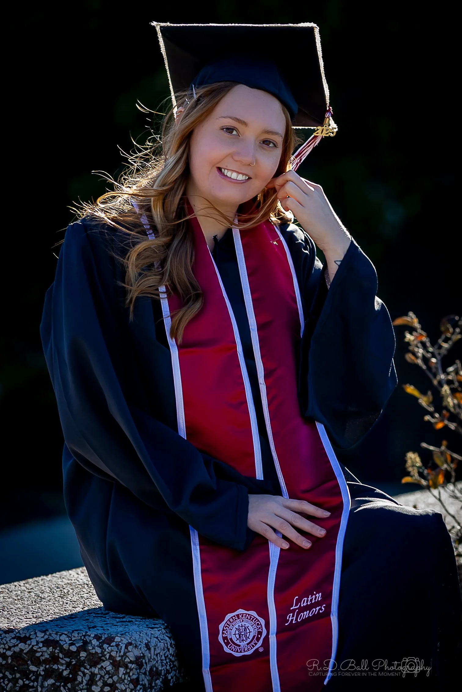 Young woman in black graduation gown and cap, sitting outdoors on a bench, holding a tassel, with a bright smile, during her graduation ceremony.
