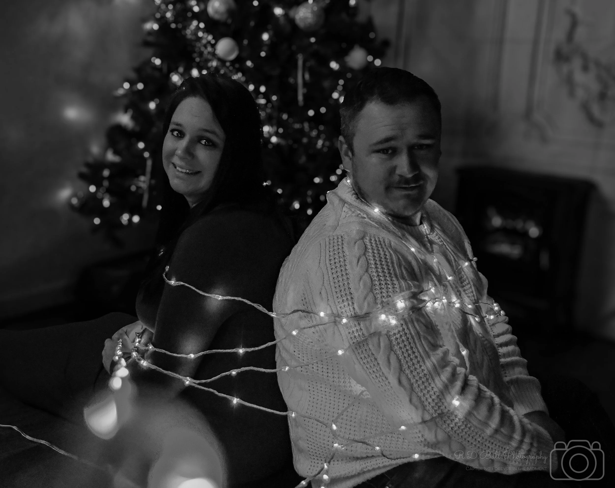 A man and woman sit back to back, surrounded by Christmas lights, in front of a decorated Christmas tree in a cozy room.