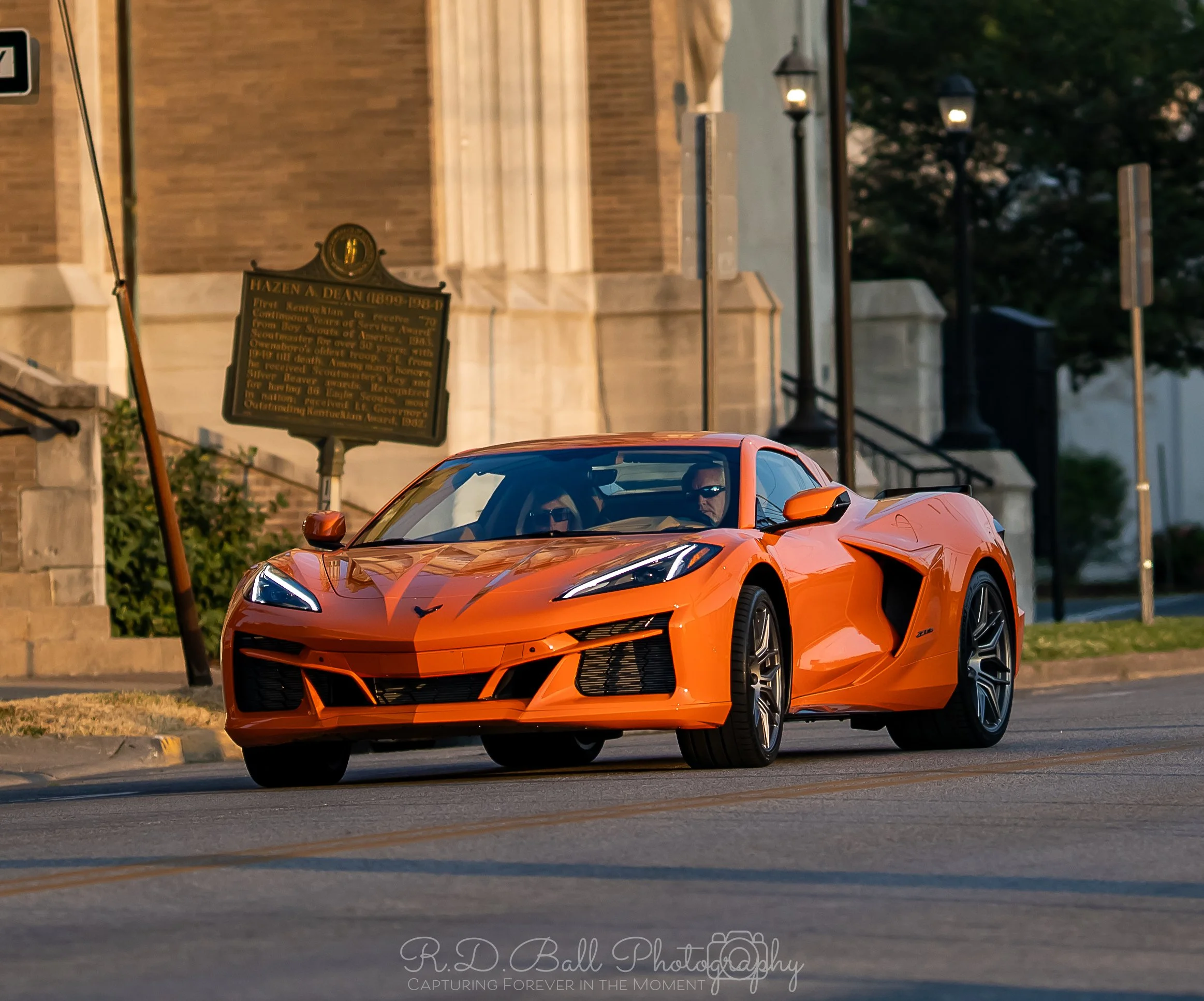 An orange Chevrolet Corvette sports car driving on a city street, with a historical marker and buildings in the background.