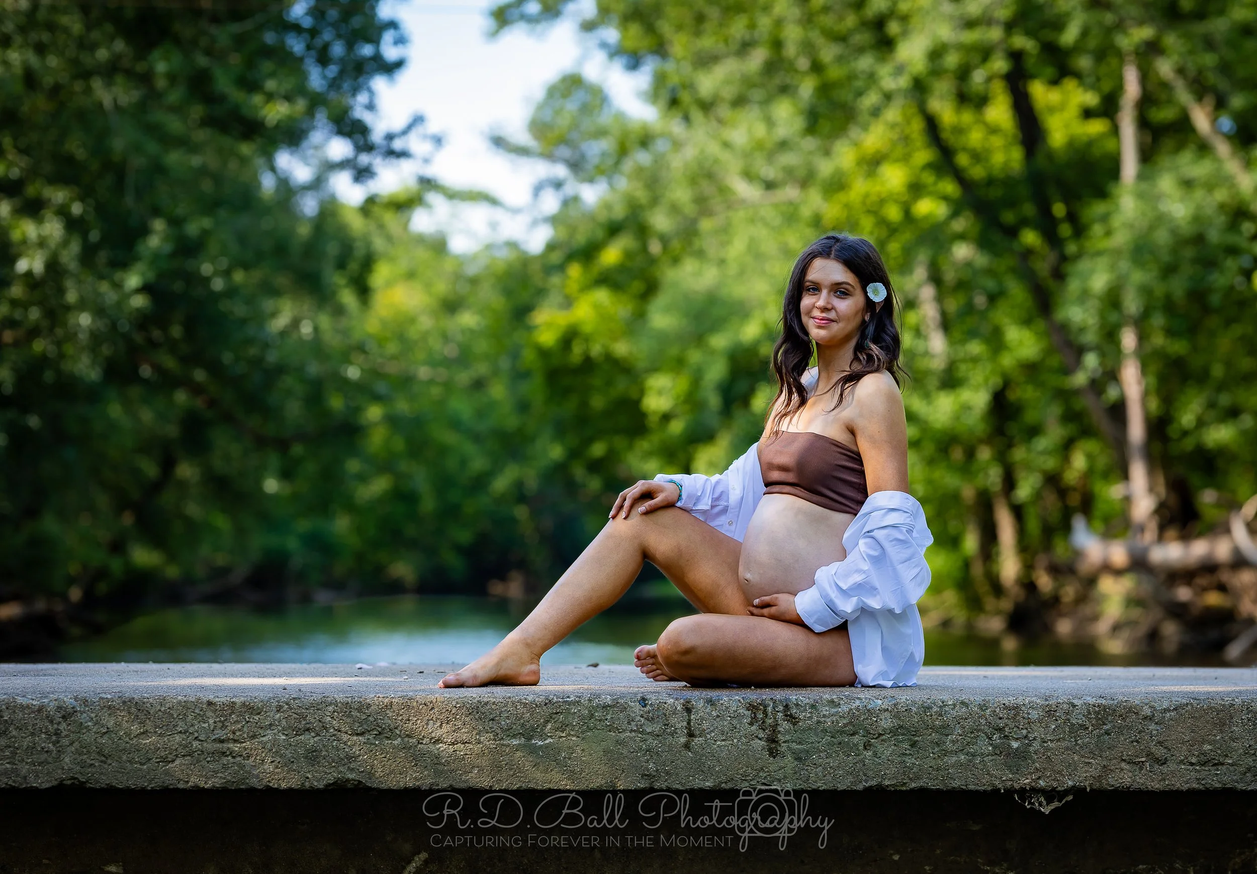 A pregnant woman sitting on a concrete surface outdoors, surrounded by green trees and water, wearing a brown tube top and white open shirt, with a flower in her hair.