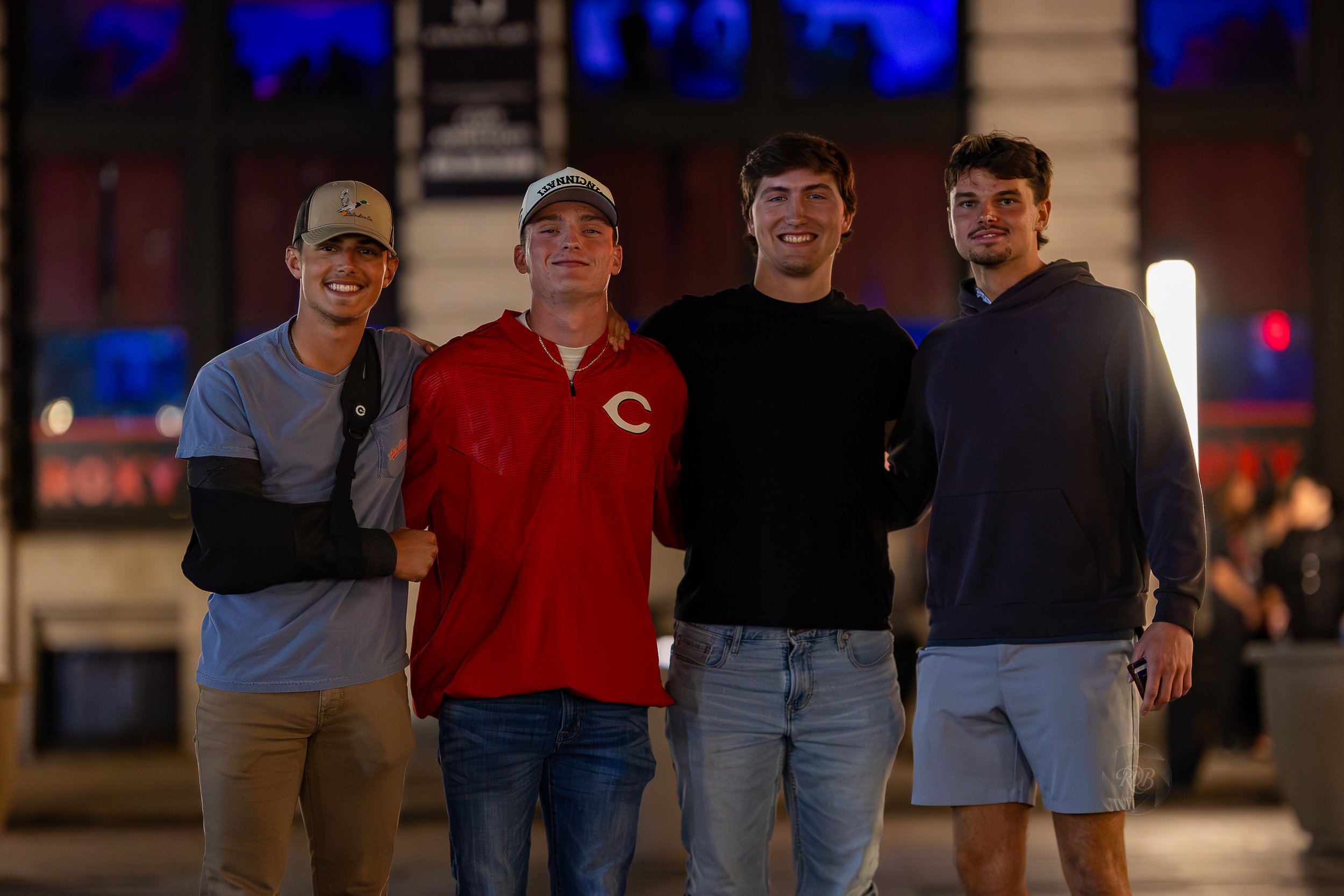 Four young men standing together outdoors at night, smiling and posing for the photo, with a blurred background showing lights and people.