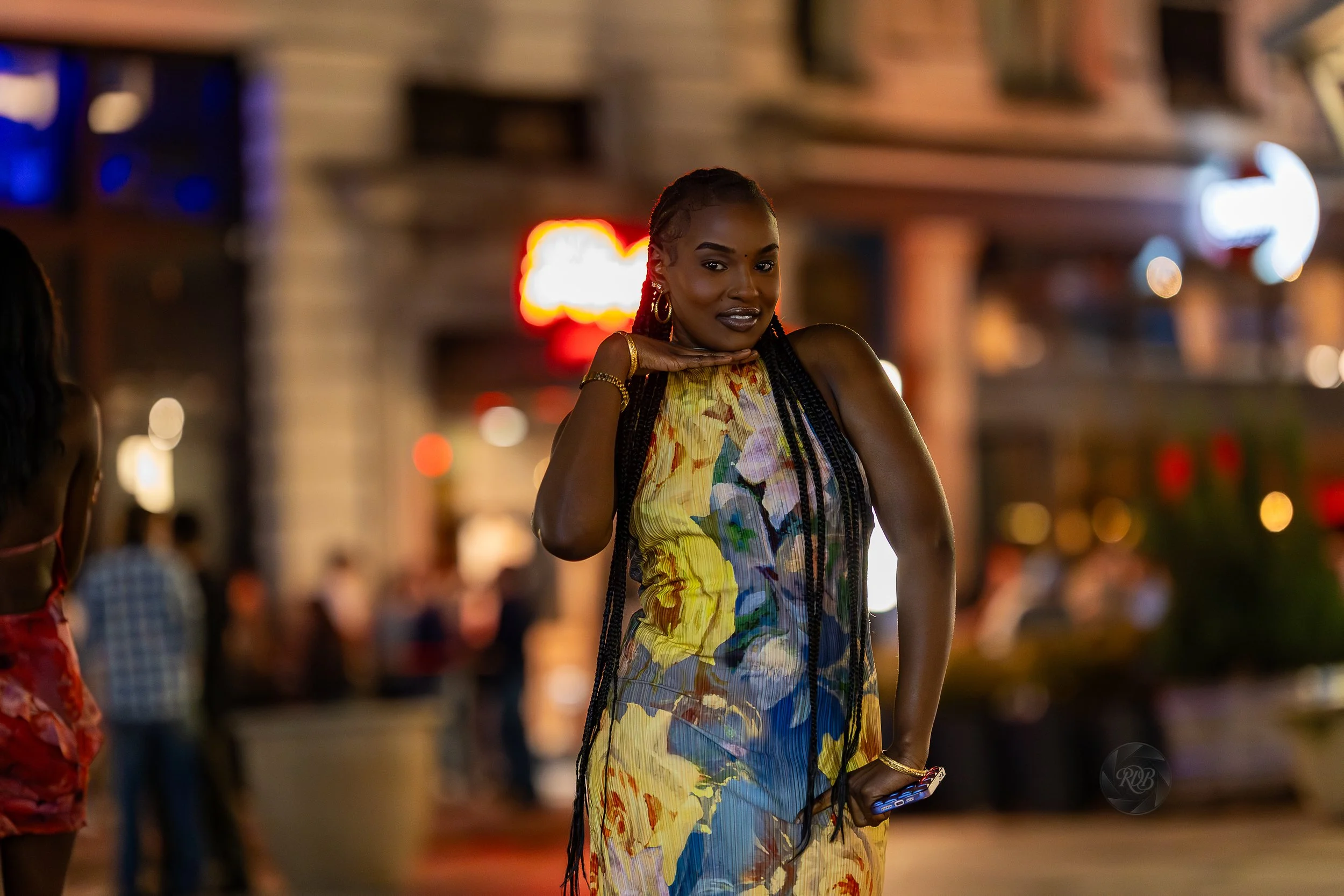 A woman in a colorful, sleeveless dress posing with her hand under her chin in a lively street scene at night, with blurred lights and people in the background.