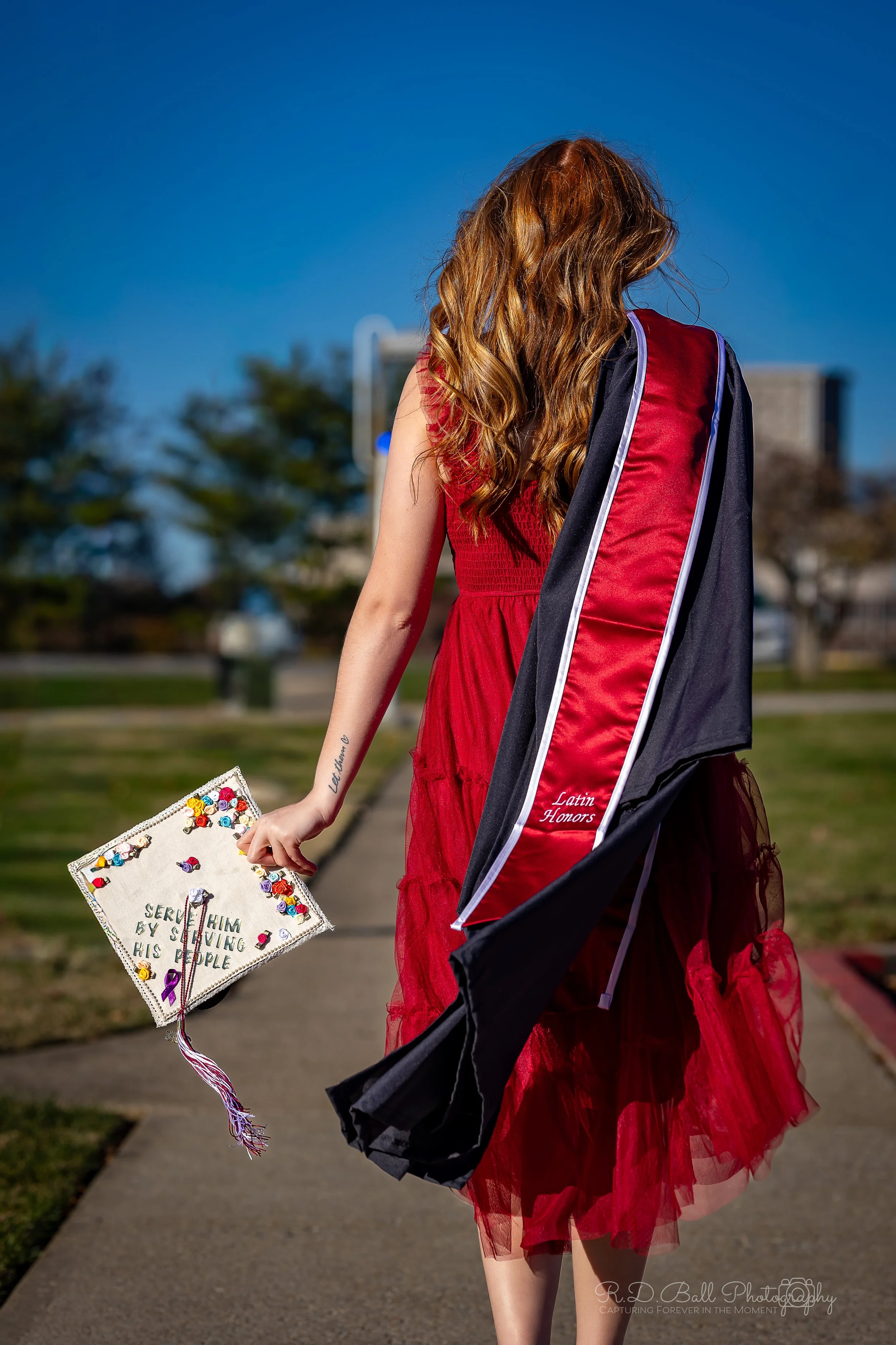 A young woman in a red dress walks on a sidewalk carrying a decorated graduation cap and a rolled-up graduation gown draped over her shoulder.