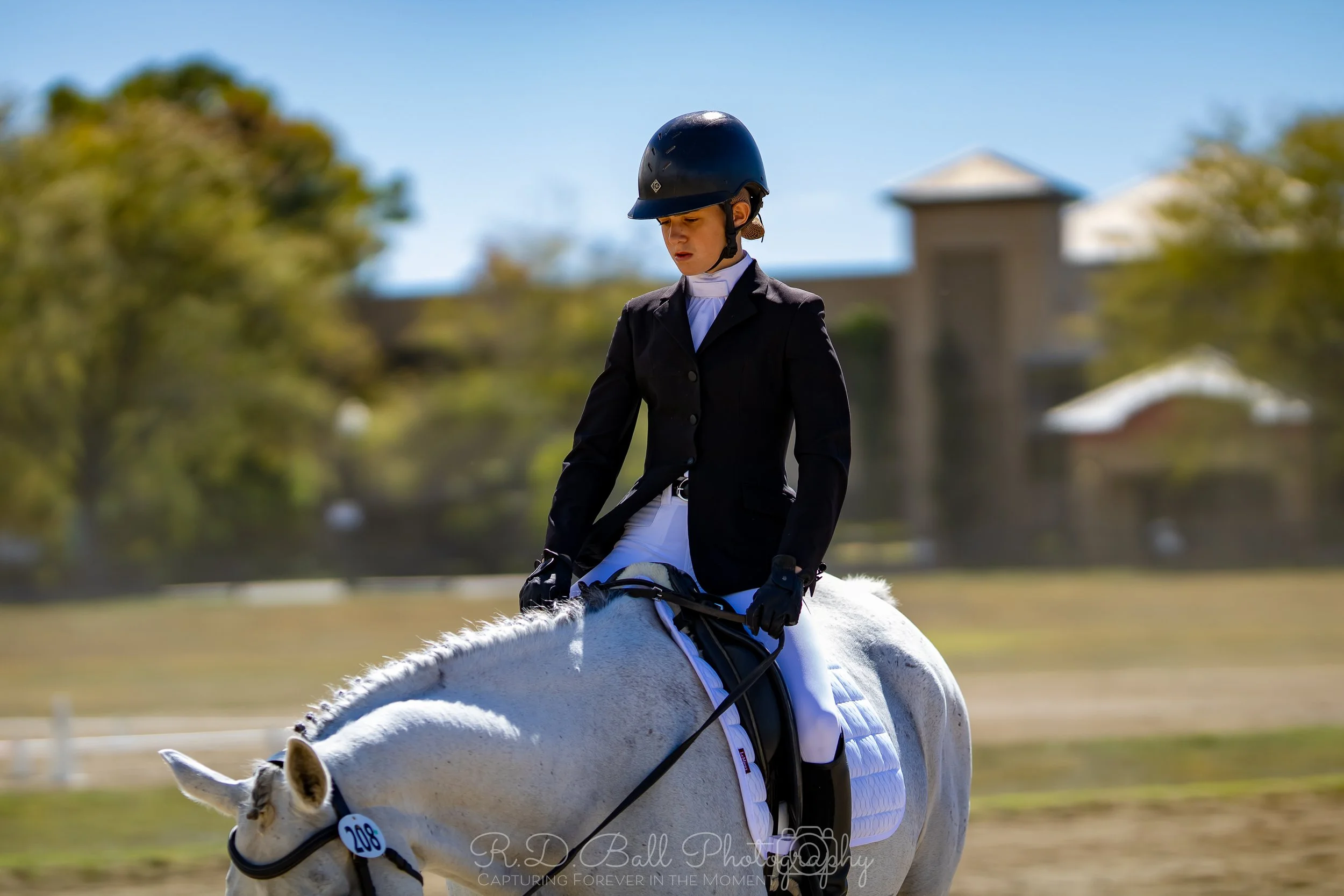 Young female equestrian rider in formal riding attire and helmet riding a white horse in an outdoor arena.