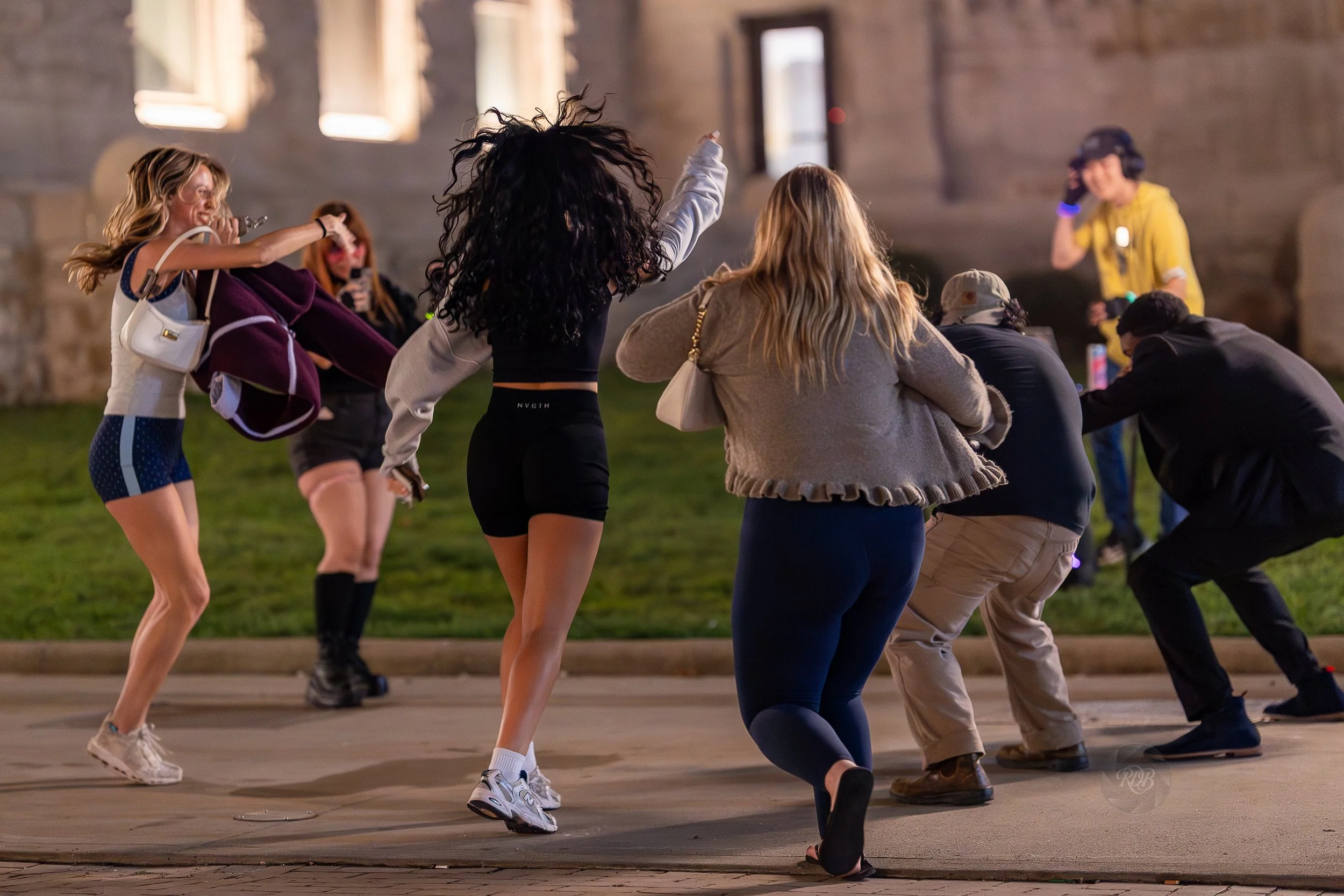 A group of people dancing outdoors at night in front of a stone building with windows, with a person in a yellow shirt in the background.