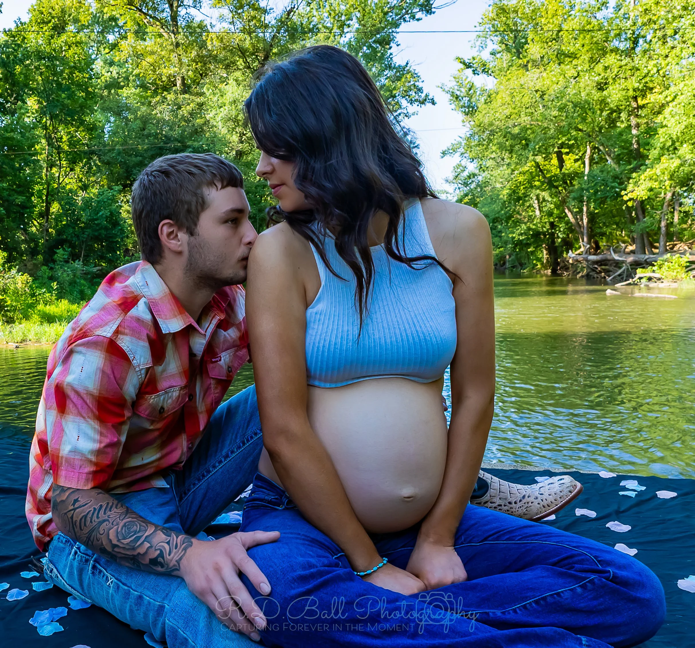 A young man and a pregnant woman sitting on a blanket by a river, with the man kissing the woman's shoulder. The woman is wearing a light blue crop top and jeans, while the man is in a red plaid shirt and jeans. There are flower petals on the blanket