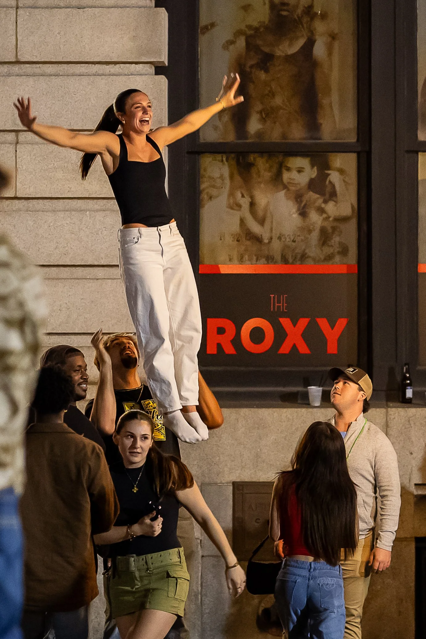 A woman in black top and white pants is being lifted by friends outside the Roxy theater, smiling with her arms outstretched. Others watch and smile, with a couple of friends looking up at her.
