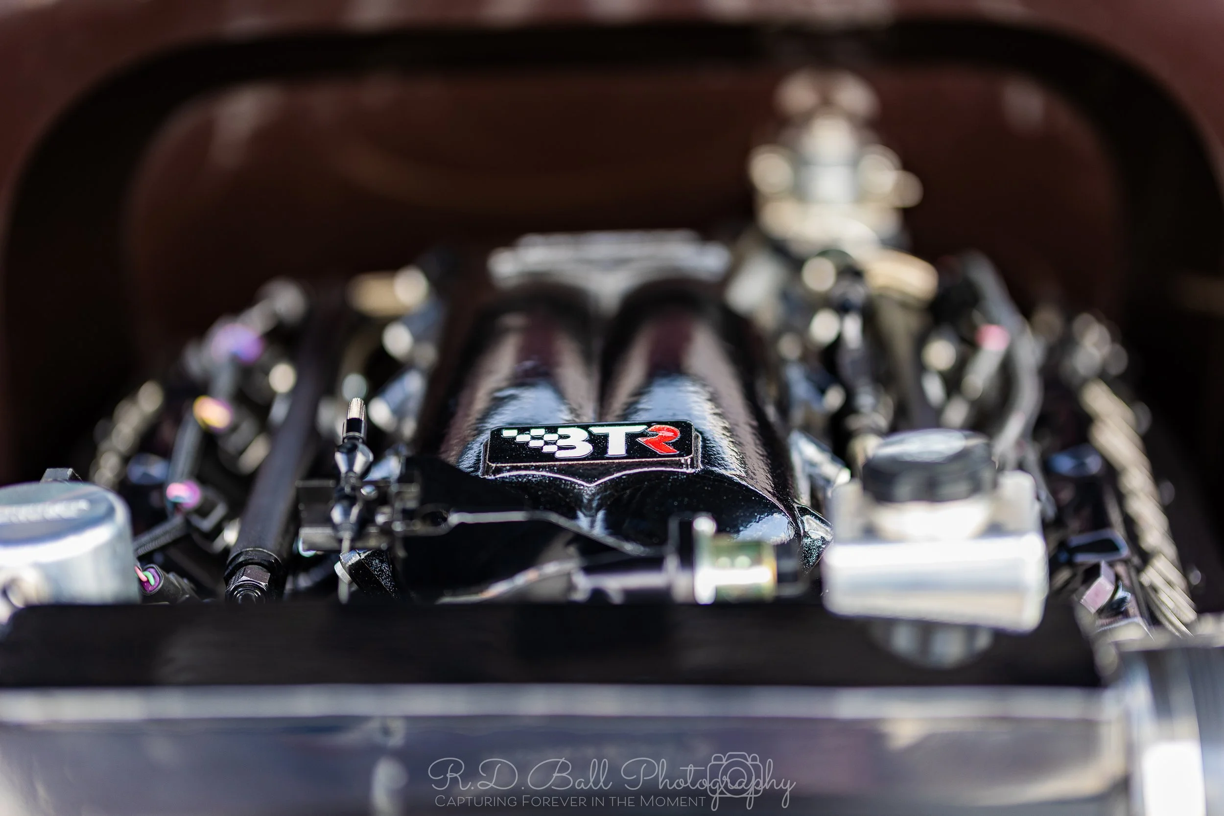 Close-up of a high-performance car engine, prominently featuring a black intake manifold with a logo that reads '3TR' in red and white. The engine is shiny and well-maintained, with various metallic components surrounding it.