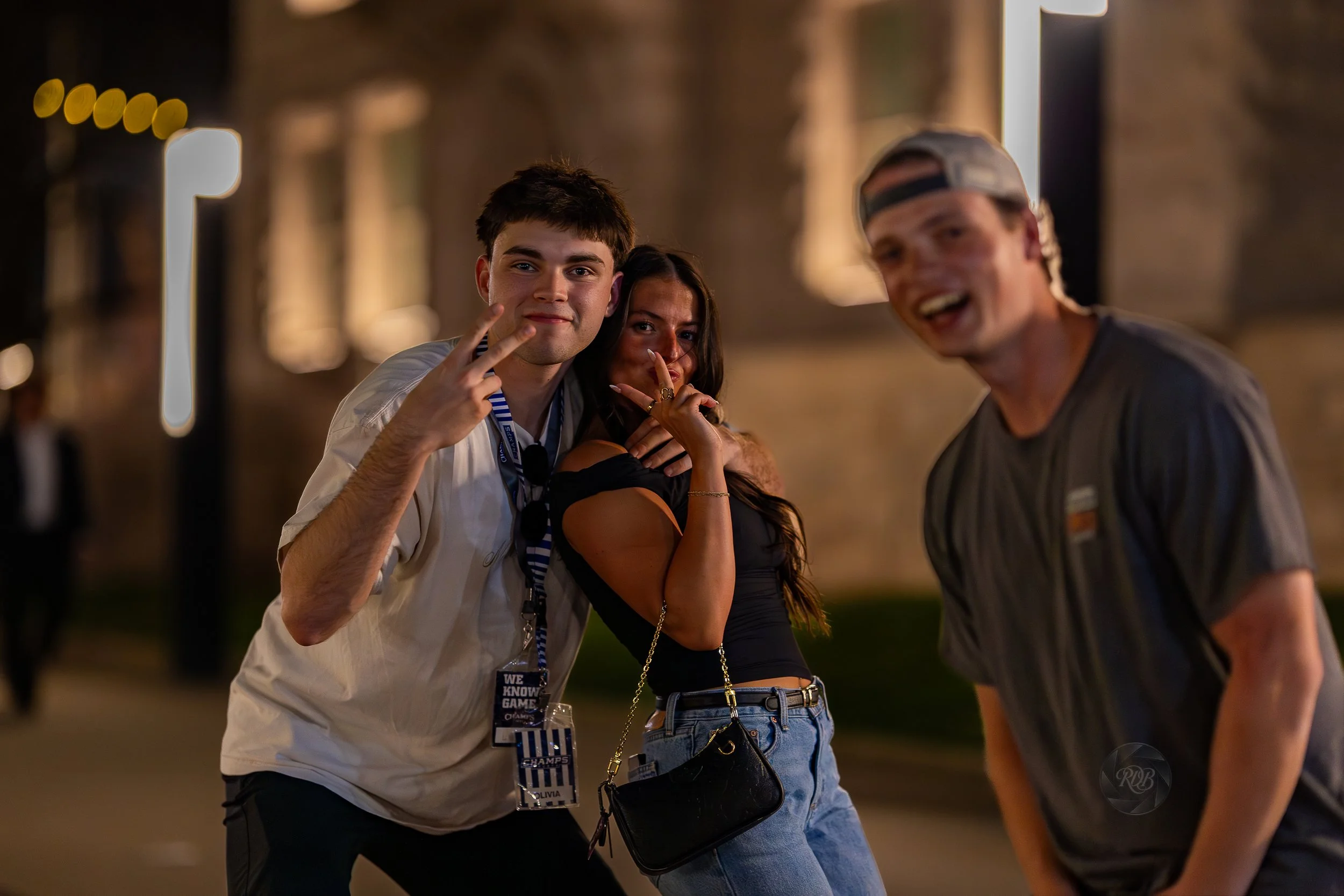 Three young adults smiling and posing for a photo at night outdoors. Two of them are giving peace signs, and they are standing close together with a blurred person in the background.