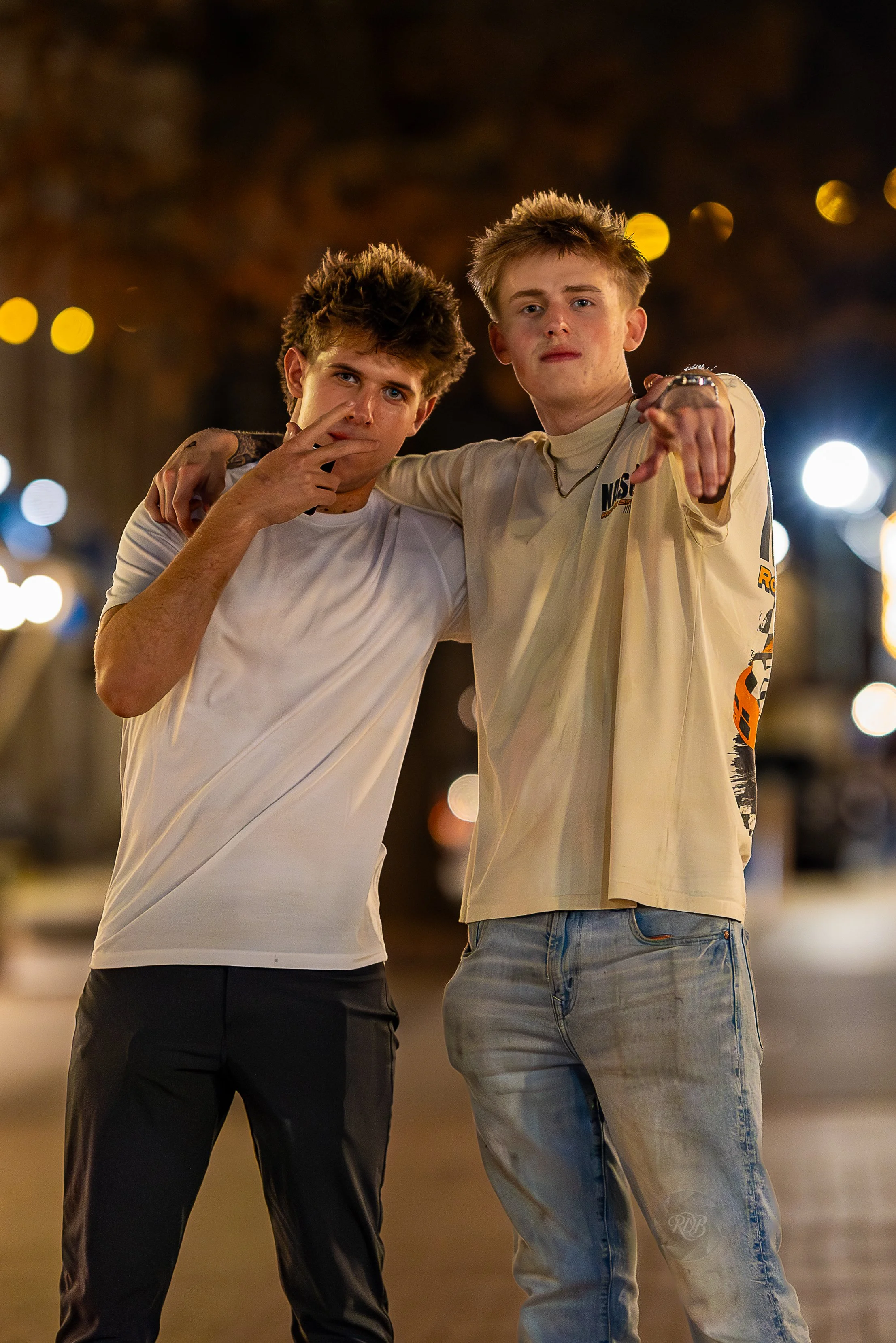Two young men standing closely together on a city street at night, with blurred lights in the background. One has his arm around the other's shoulder, and the one on the left has his finger near his lips. The man on the right is pointing towards the 