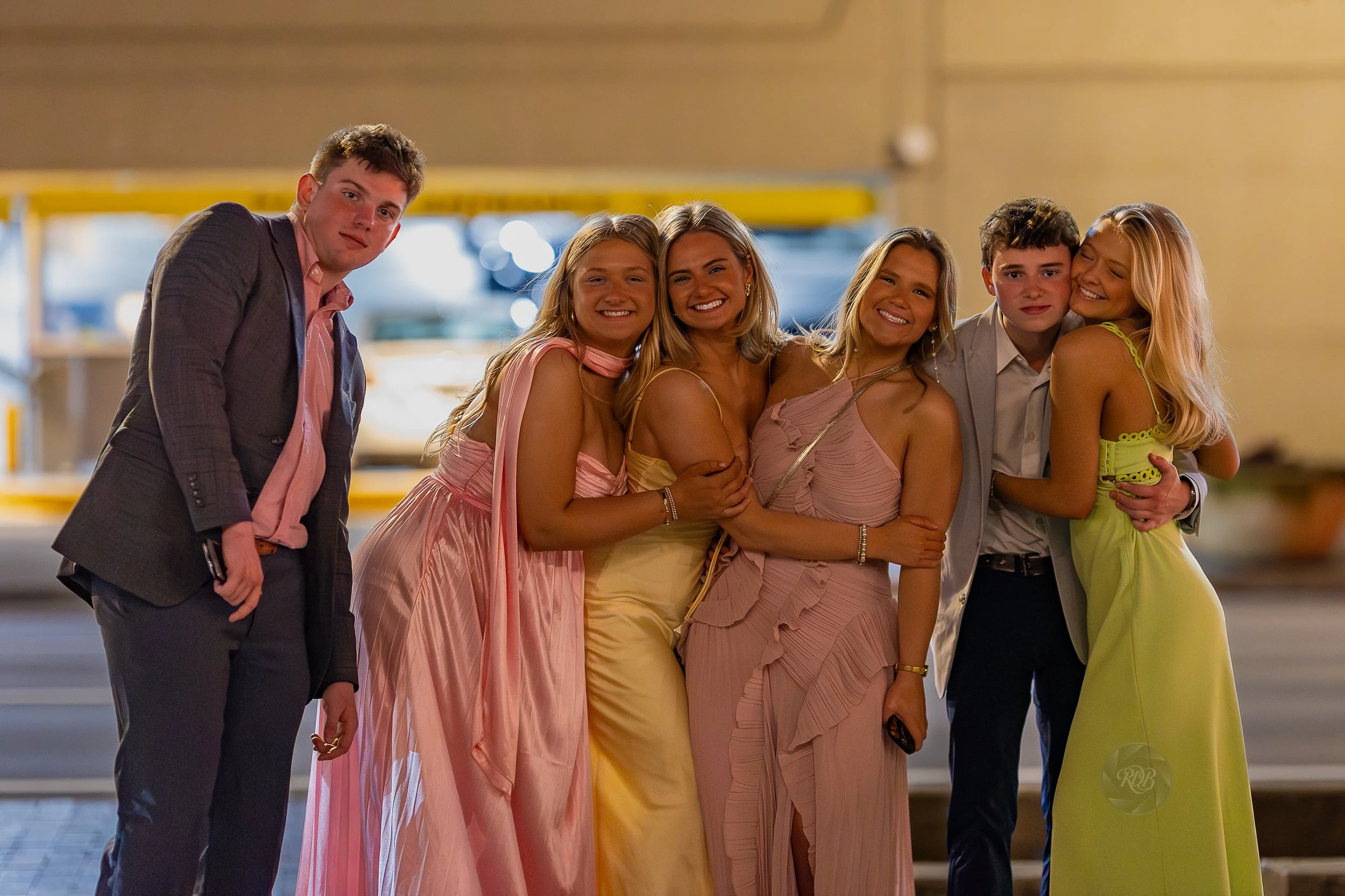 Group of six young people dressed in formal attire, standing close together and smiling, at nighttime outdoor setting.