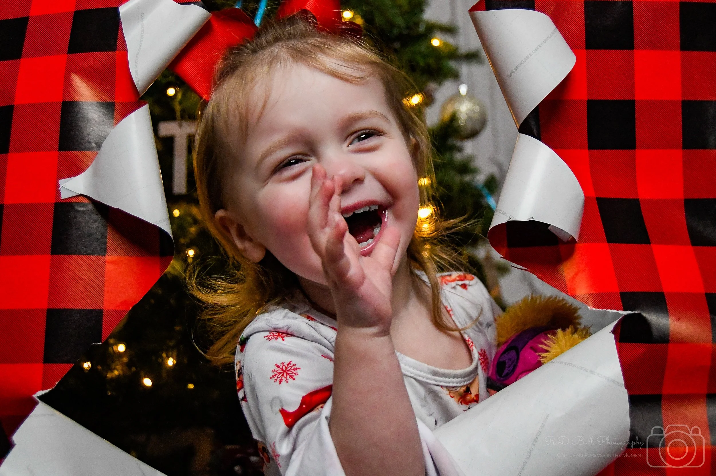 A young girl with red hair and festive pajamas is smiling and laughing while sitting in a holiday wrapping paper cutout, with a Christmas tree decorated with lights and ornaments in the background.