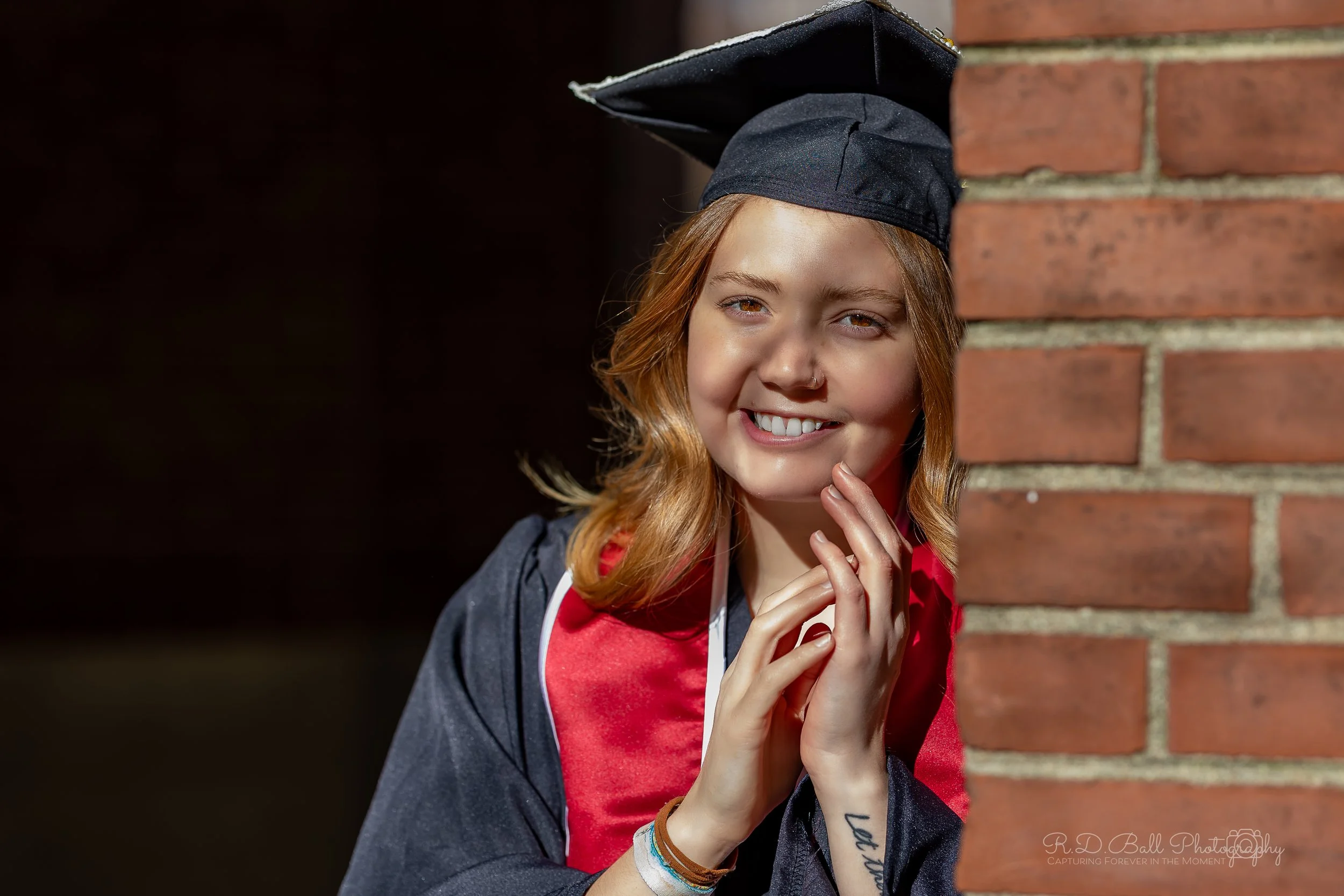 Young woman in graduation cap and gown smiling, leaning on a brick wall.