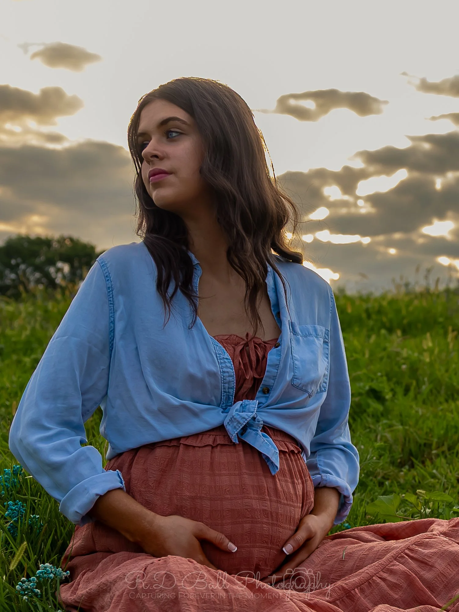 A pregnant woman sitting outdoors in a field during sunset, wearing a blue tied-up shirt over a rust-colored dress, with her hands resting on her belly.