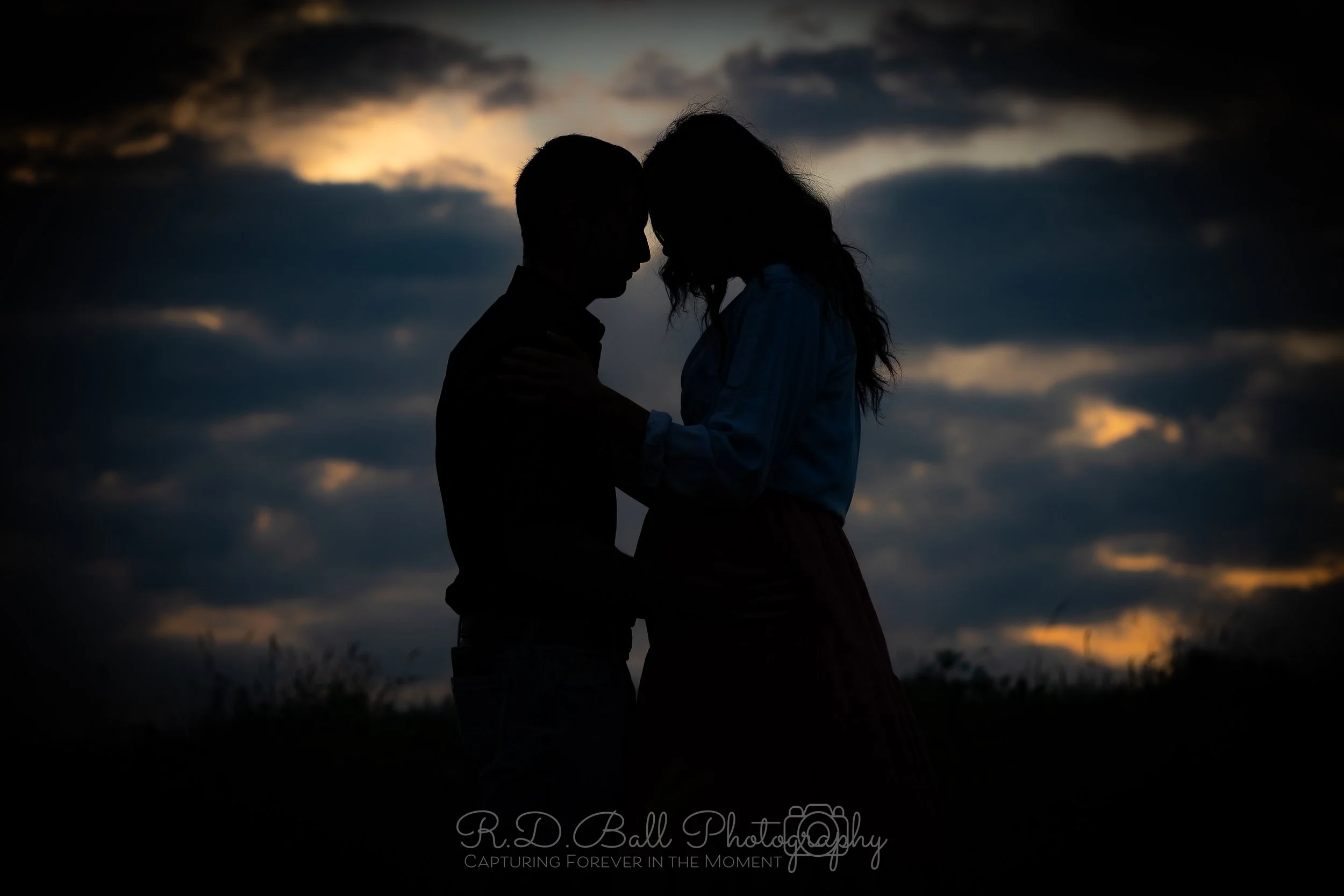 Silhouette of a couple embracing outdoors at sunset, with dark clouds in the sky.