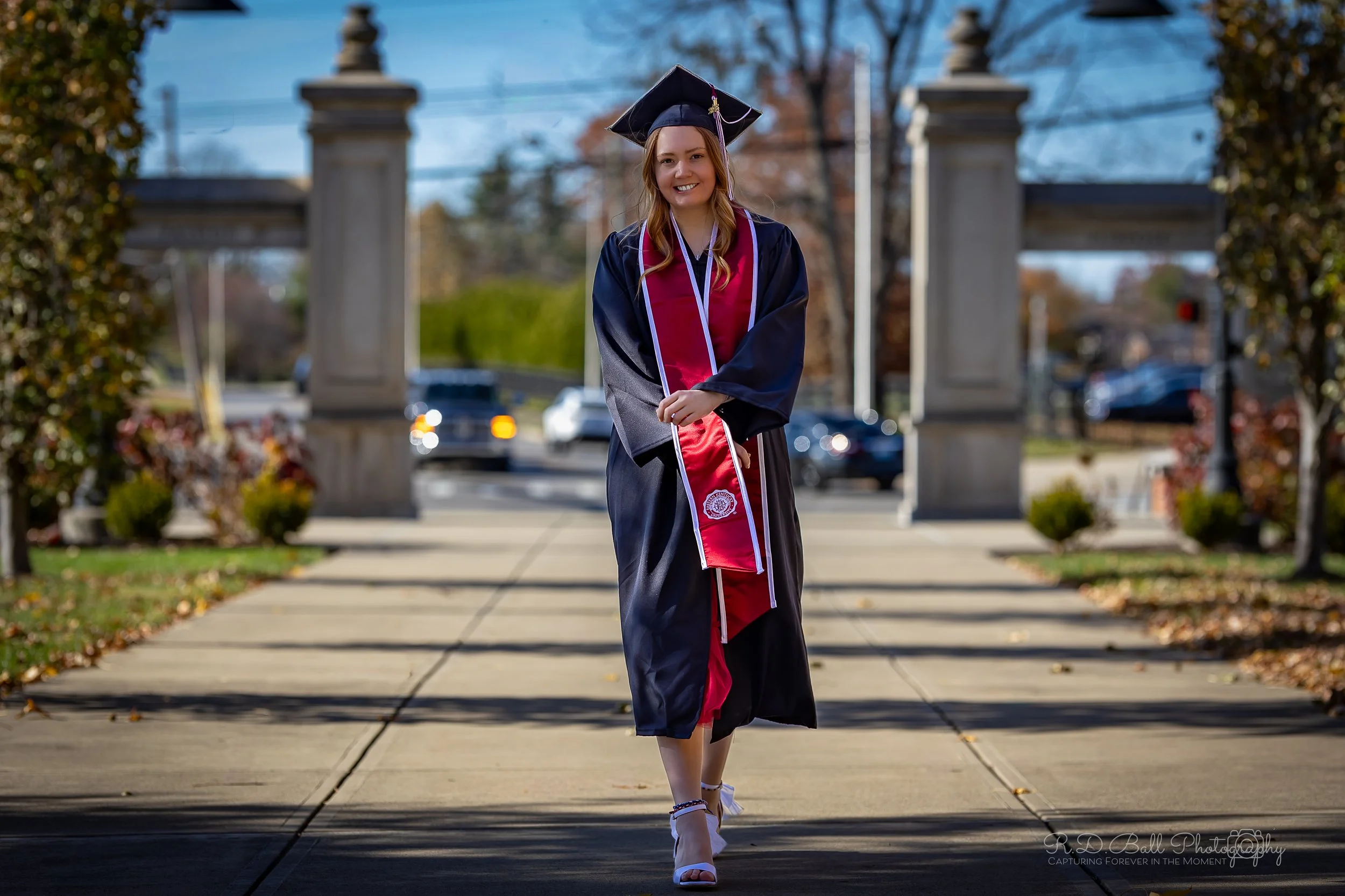 A young woman in a graduation cap and gown walking outdoors on a sidewalk, smiling, with trees and an archway in the background.