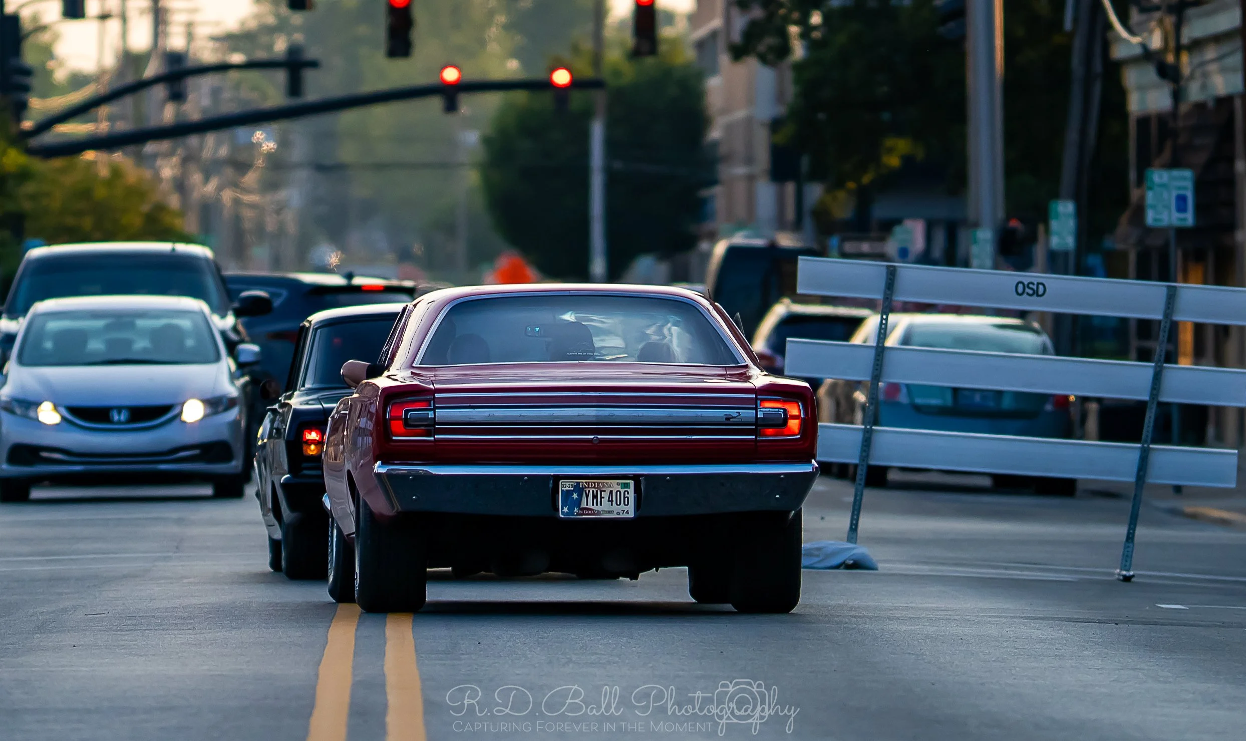 A red vintage car stopped in the middle of a city street with a temporary barricade blocking part of the road, and several other cars lined up behind it, with traffic lights overhead.