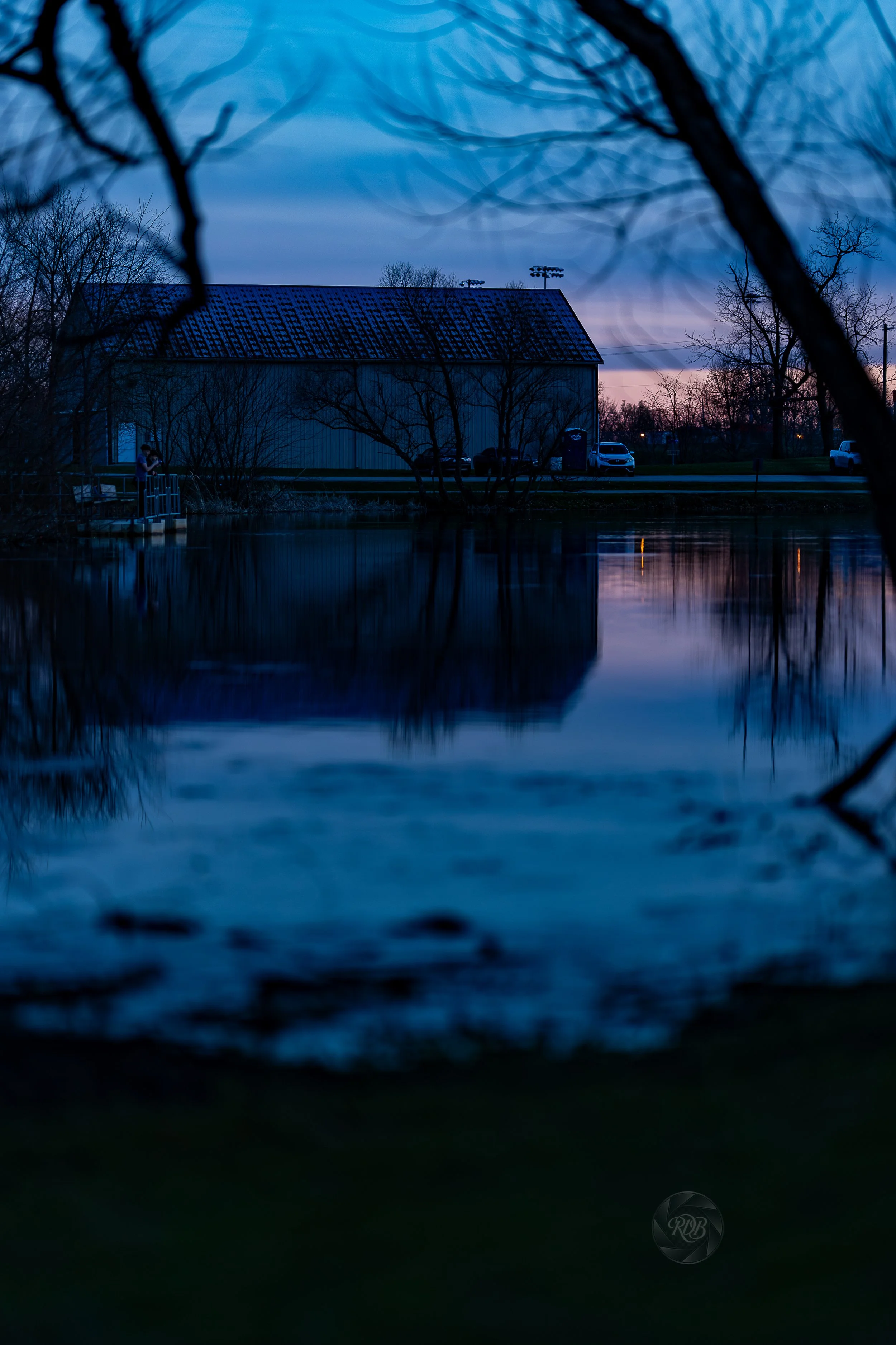 A peaceful evening scene at a pond with a barn and parked cars in the background, reflected in the water, with leafless trees and the sky transitioning from sunset to night.